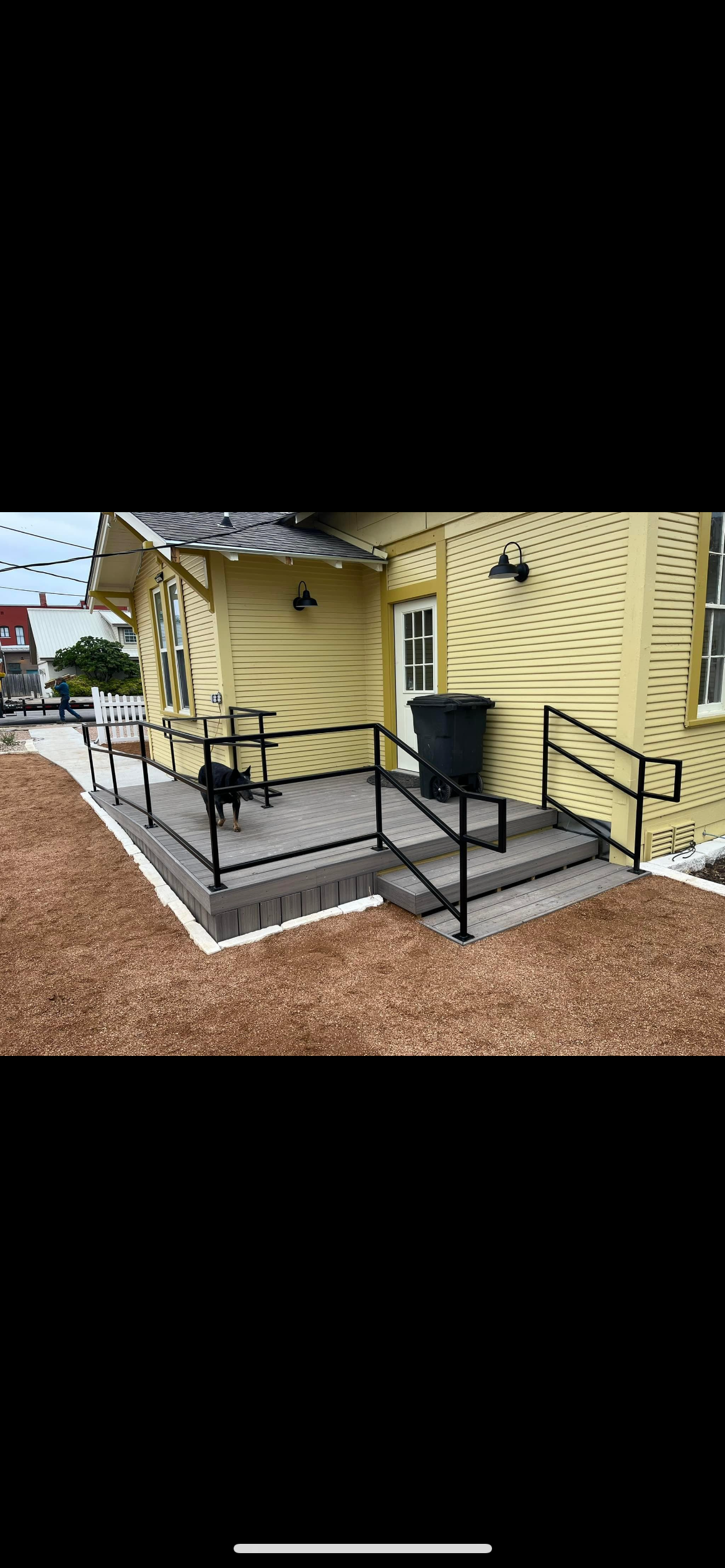 Front porch with black metal railing, yellow house exterior, and a black trash bin. A dog is standing on the porch, and a person is walking in the background.
