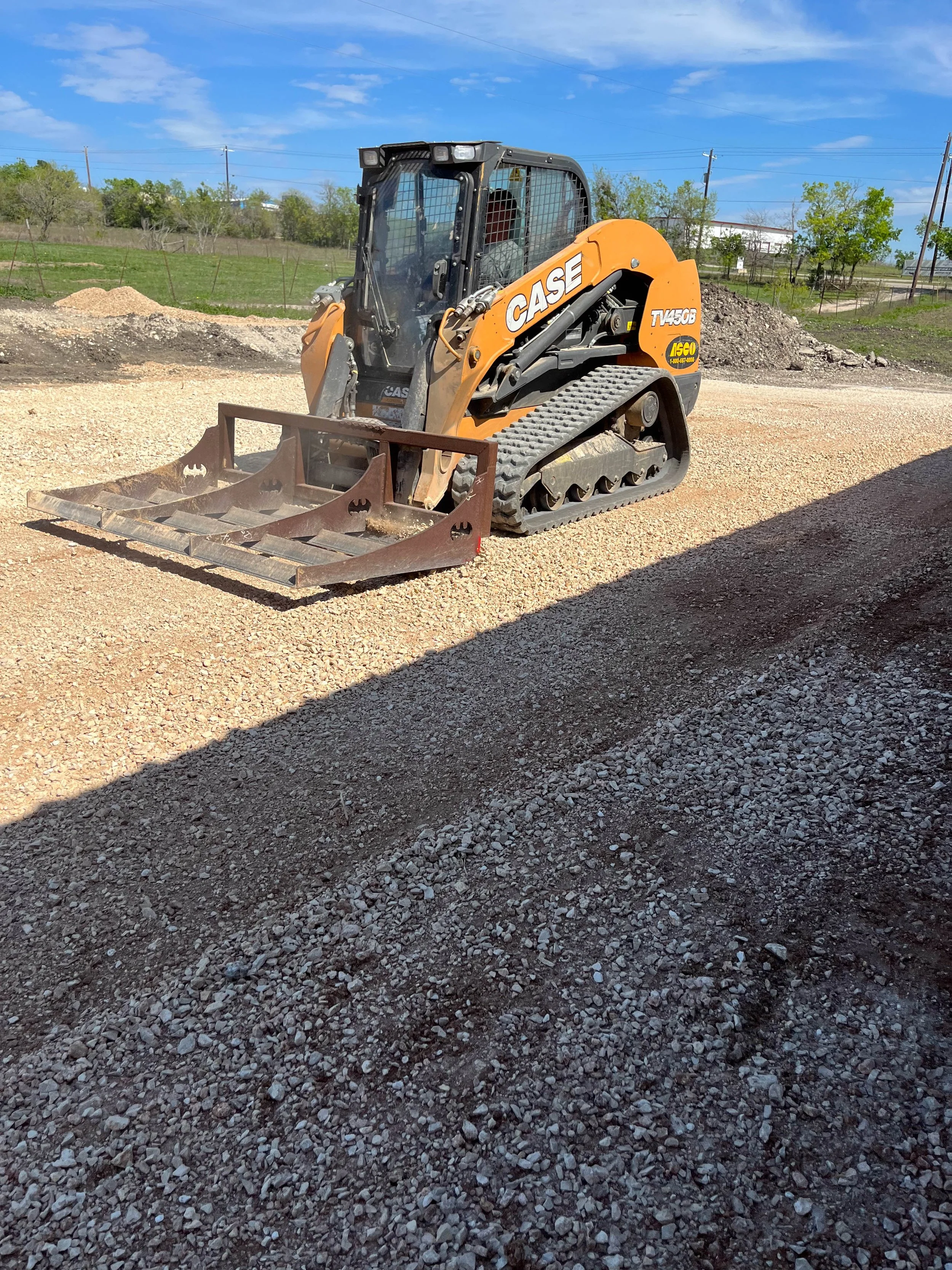 A small construction vehicle, a CASE T750B compact track loader, on a gravel surface at a construction site with dirt piles and a green landscape in the background under a blue sky with clouds.