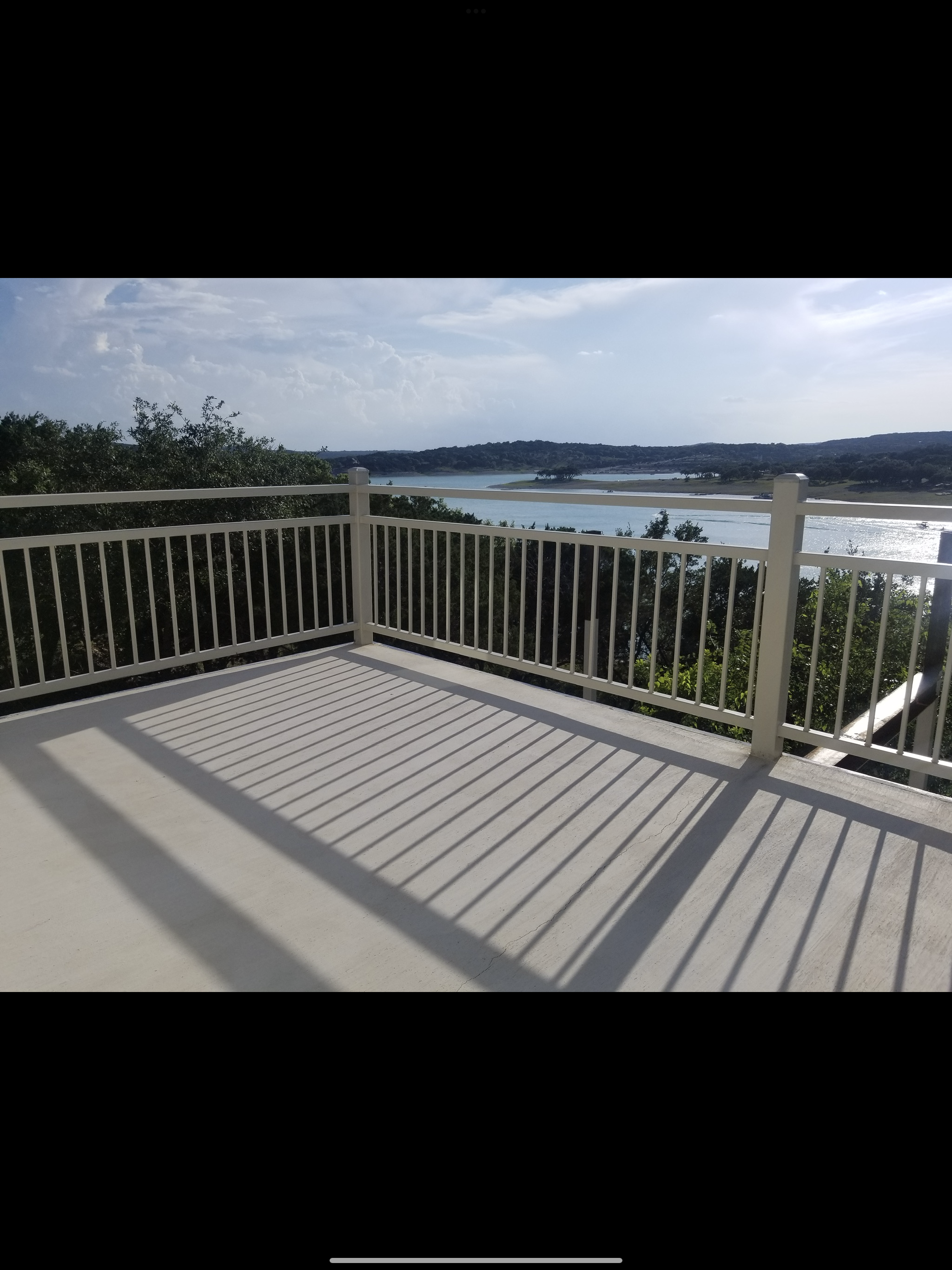 View from a balcony with a white railing overlooking a lake or reservoir with trees and hills in the distance under a partly cloudy sky.