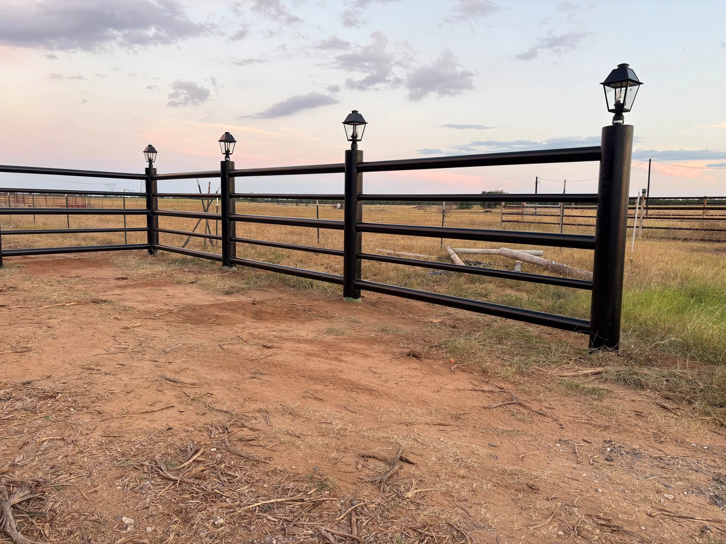 A black metal fence with decorative lamp posts in a rural area during sunset, with open fields and power lines in the background.