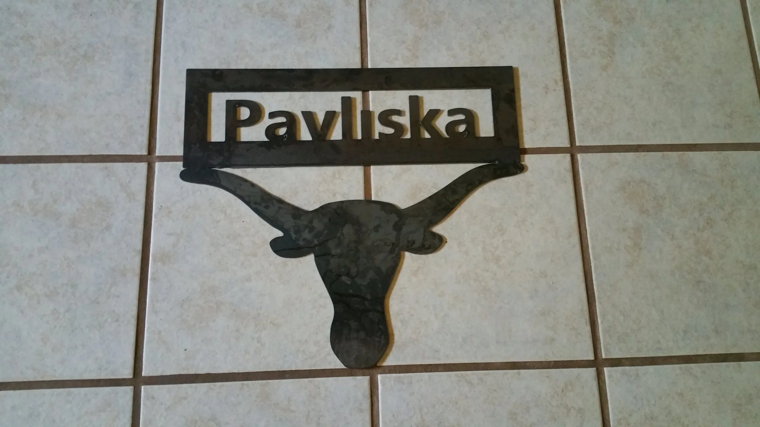 A metal wall decoration with the word 'Paviska' and a goat silhouette, mounted on a tiled wall.