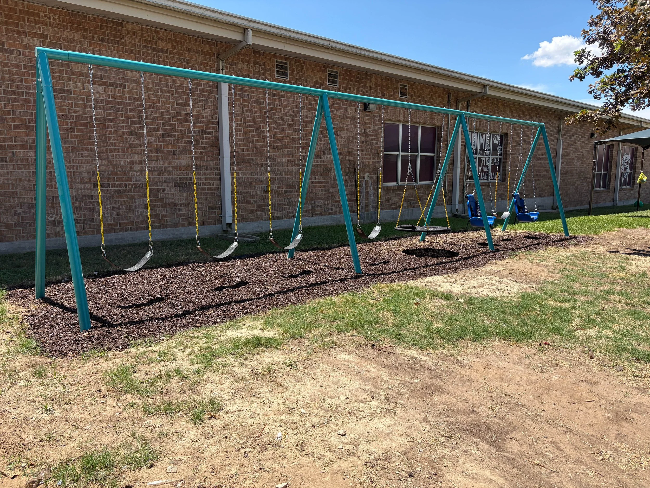 A playground swing set with four swings in front of a brick building under a partly cloudy sky.