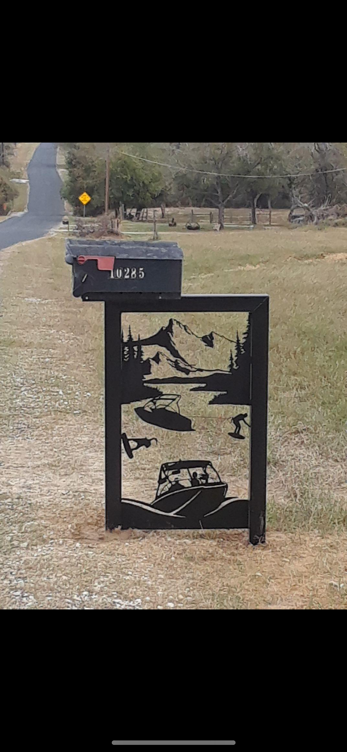 A black metal mailbox with a mounted outdoor decor panel depicting mountain scenery, trees, a lake, a boat, and two people kayaking.