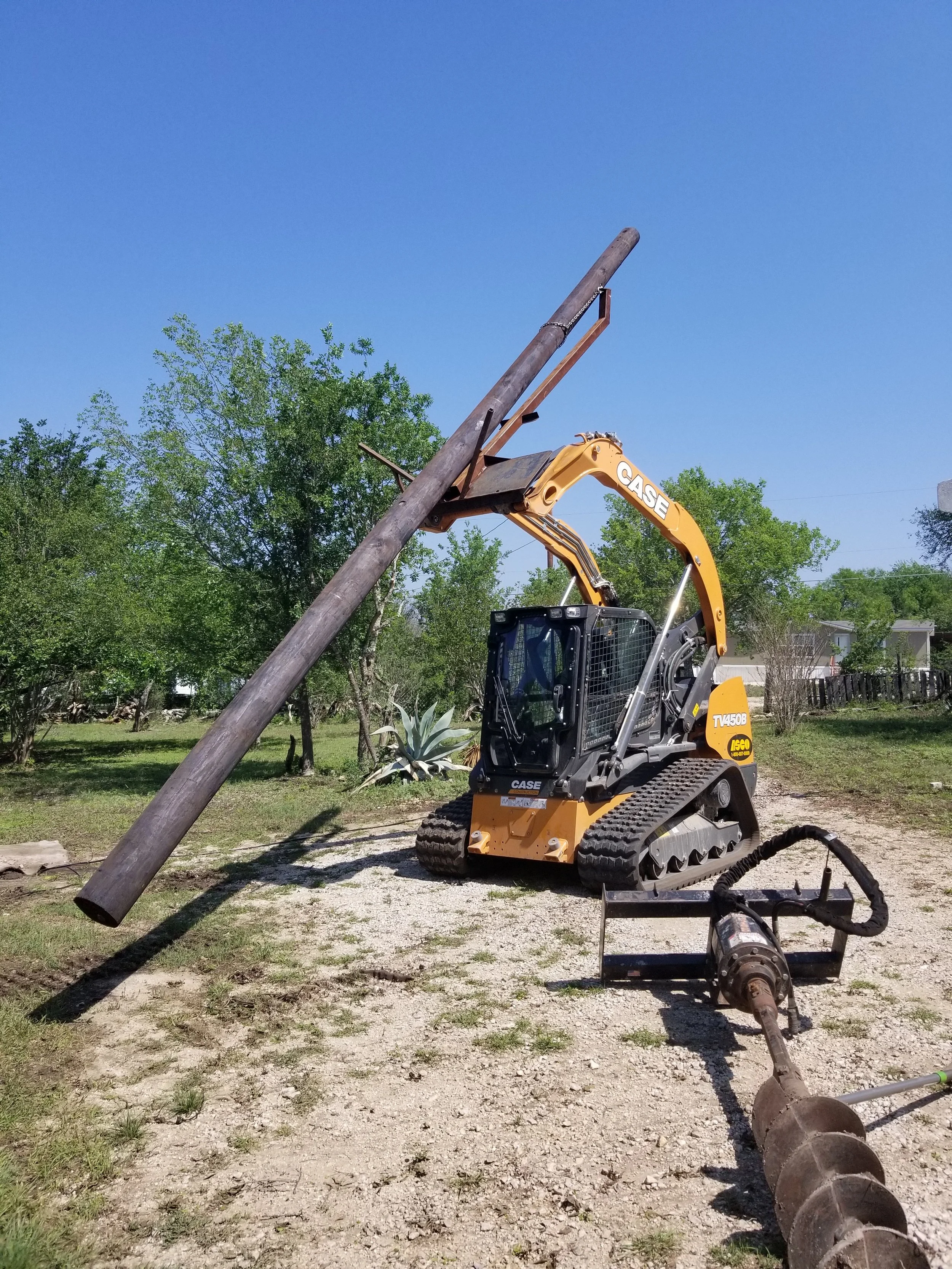 A construction site with a small CASE backhoe loader lifting a wooden utility pole. There is a large auger attachment on the ground nearby, and trees and a fence in the background.