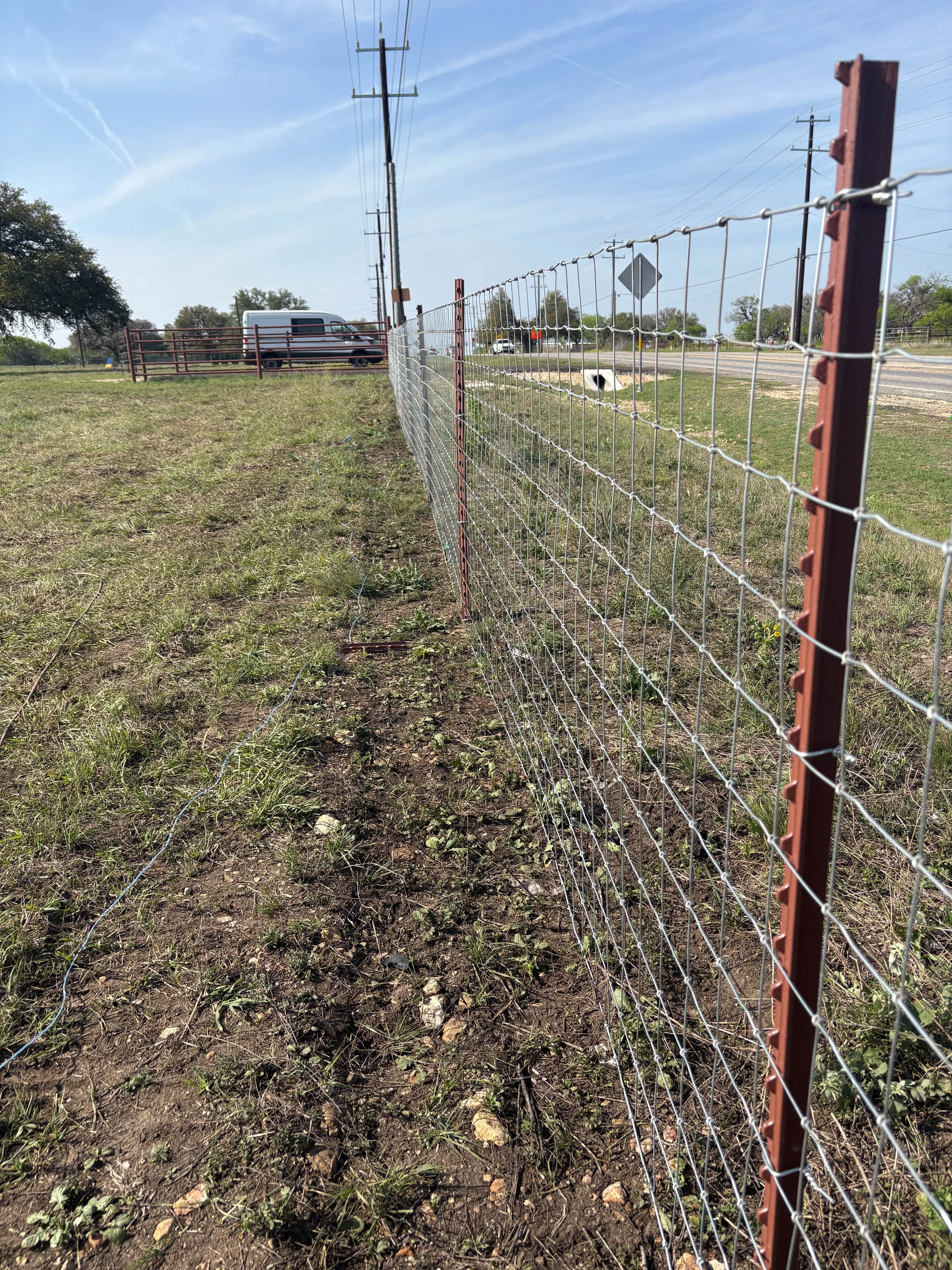 Fenced grassy area beside a road with utility poles and a white van parked near a red gate, under a partly cloudy sky.