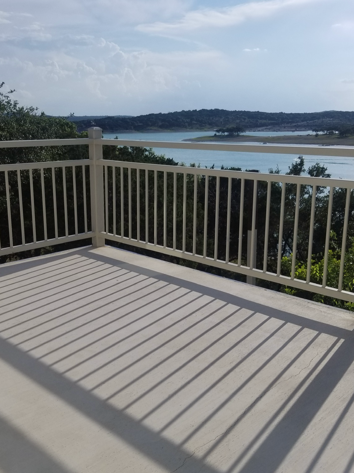 View from a balcony with a white railing overlooking a lake, trees, and distant hills under a partly cloudy sky.