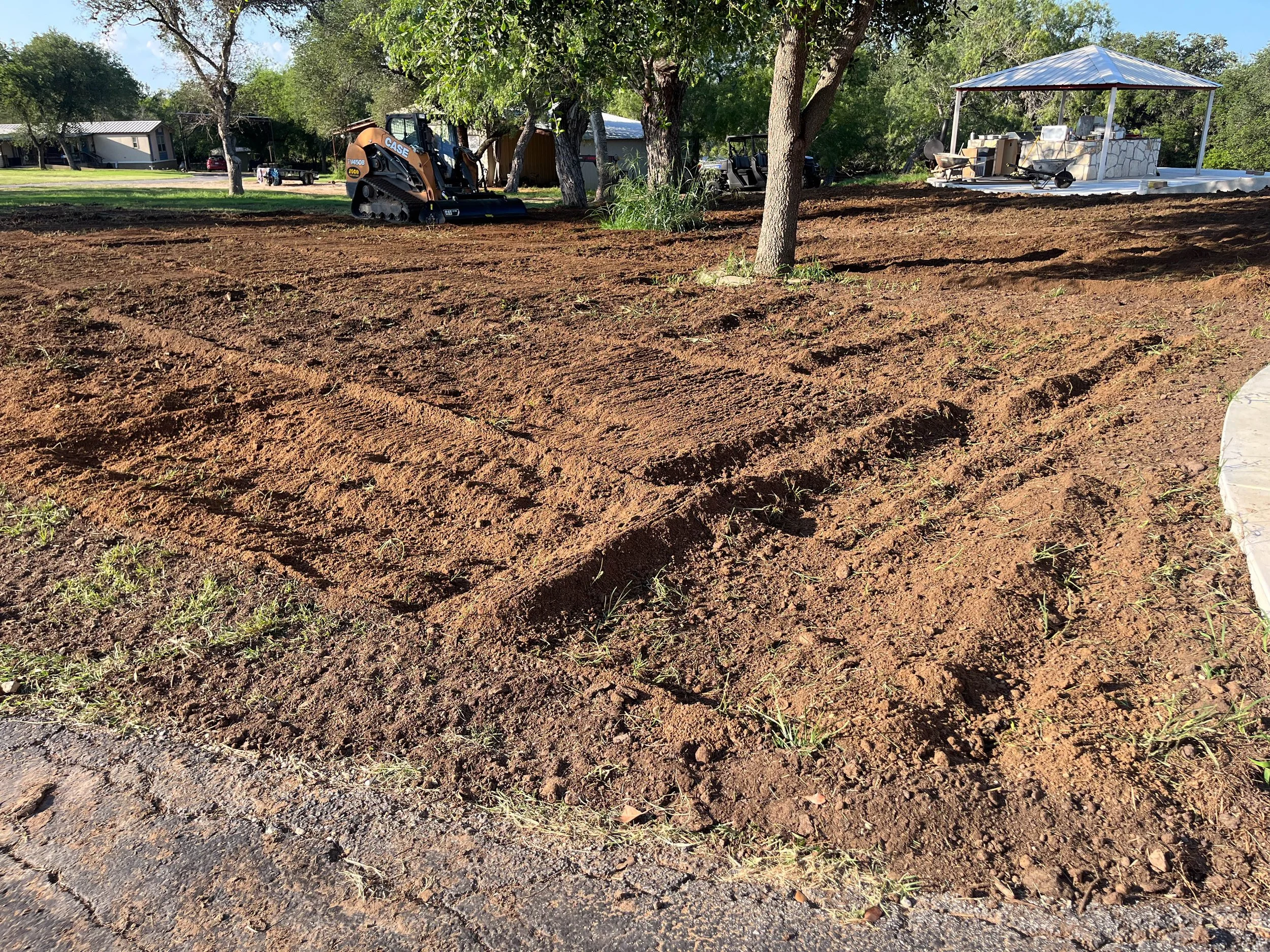 A backyard with freshly tilled soil, several trees, a small tractor, and a covered outdoor kitchen with a stone counter and cooking equipment.