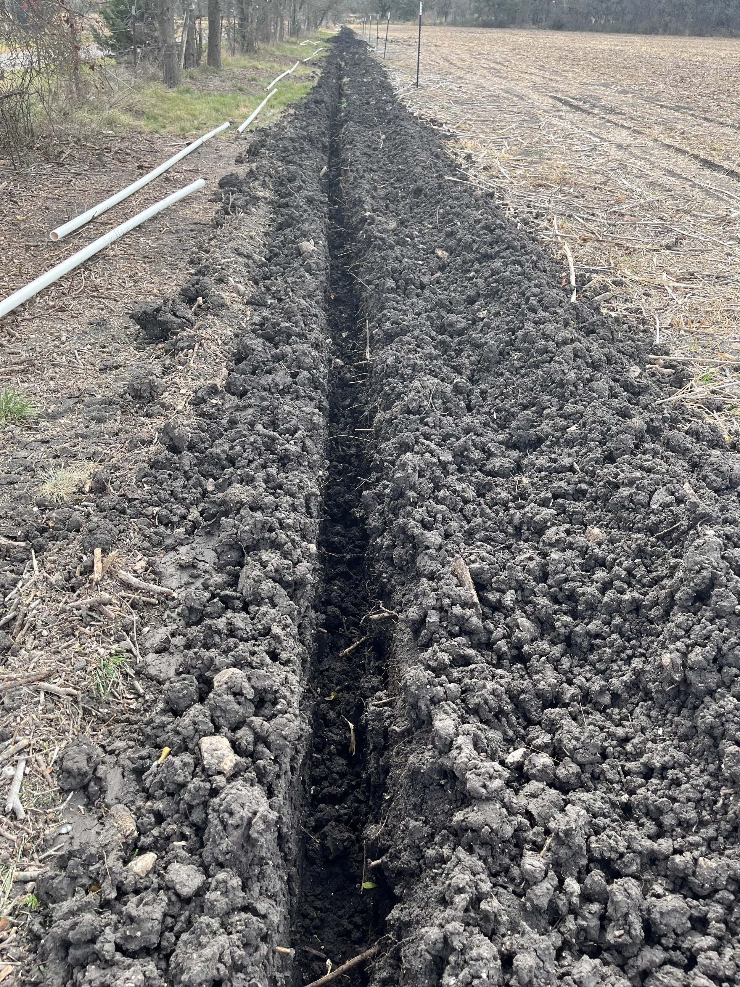 A long trench dug into the dirt alongside a field, with white PVC pipes on the ground nearby, possibly for irrigation or drainage.