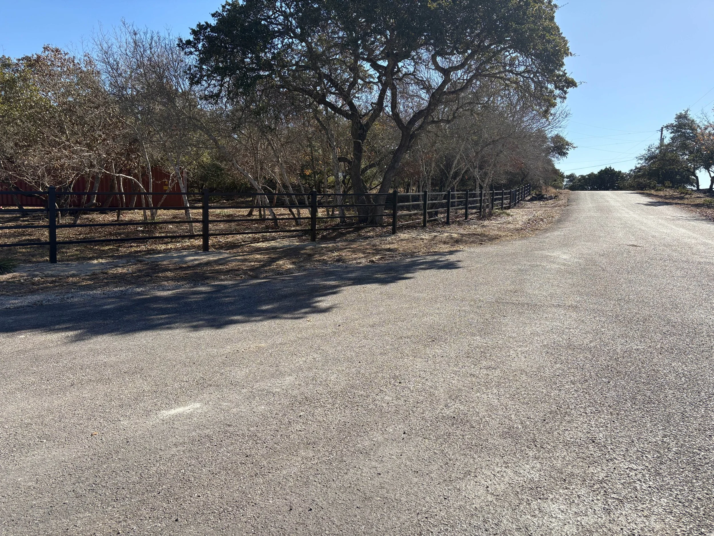 A paved country road running alongside a fence with leafless trees and some green shrubbery under a clear blue sky.
