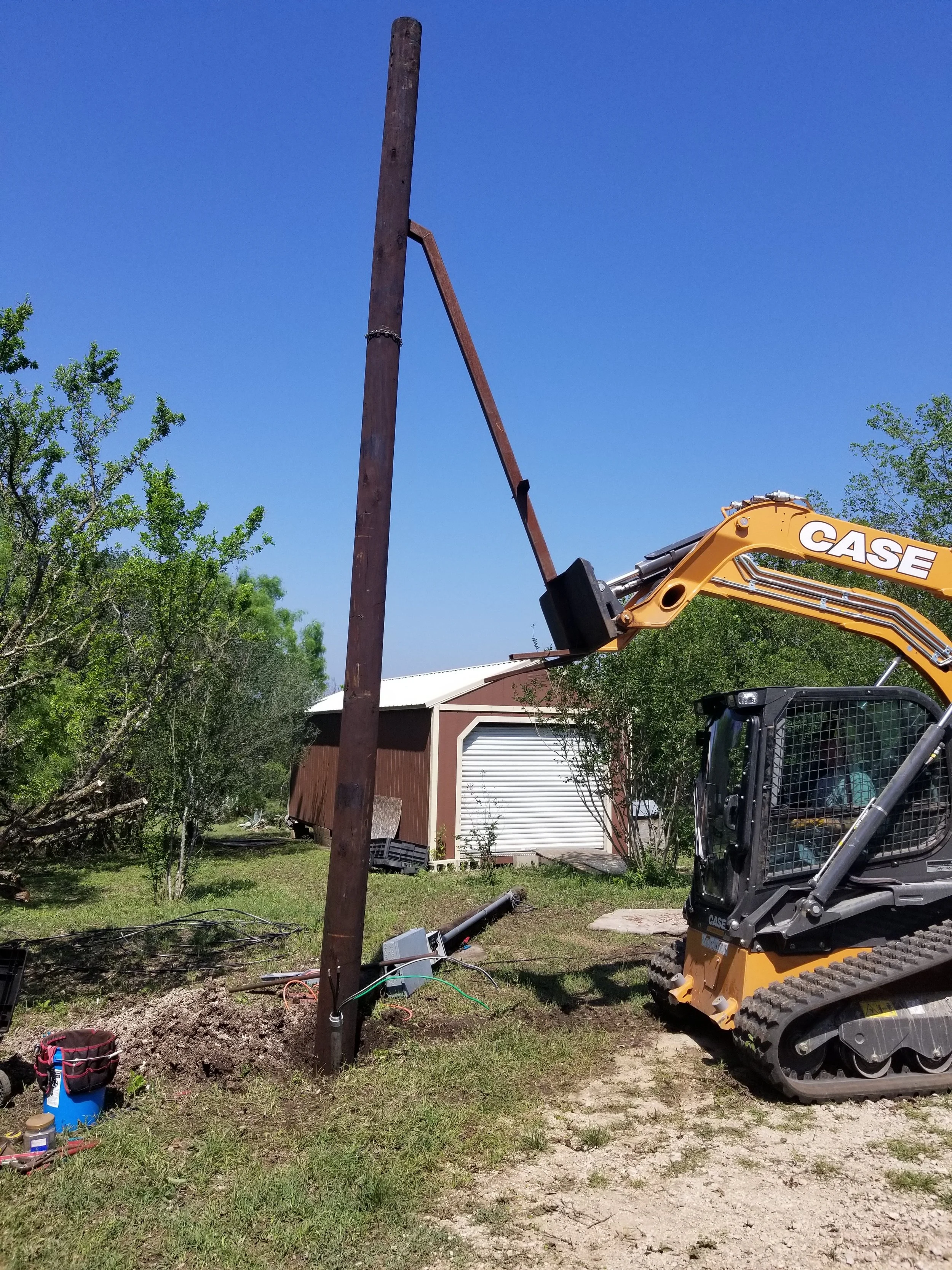 A construction scene with a mini excavator working on a utility pole installation amid a residential area with trees and a house with a white garage door under a clear blue sky.