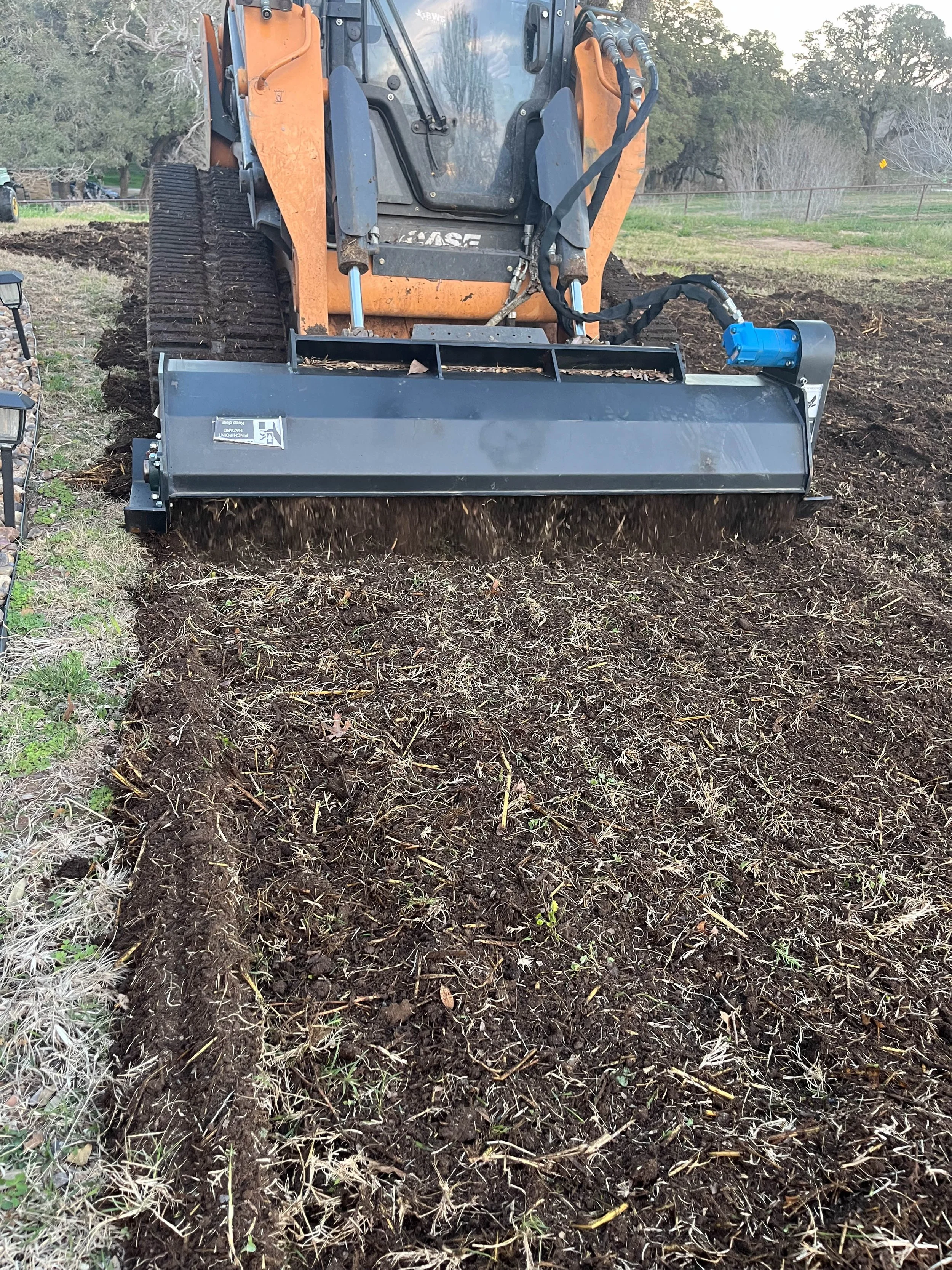 A bulldozer equipped with a hydraulic attachment working on soil, with trees and a grassy field in the background.