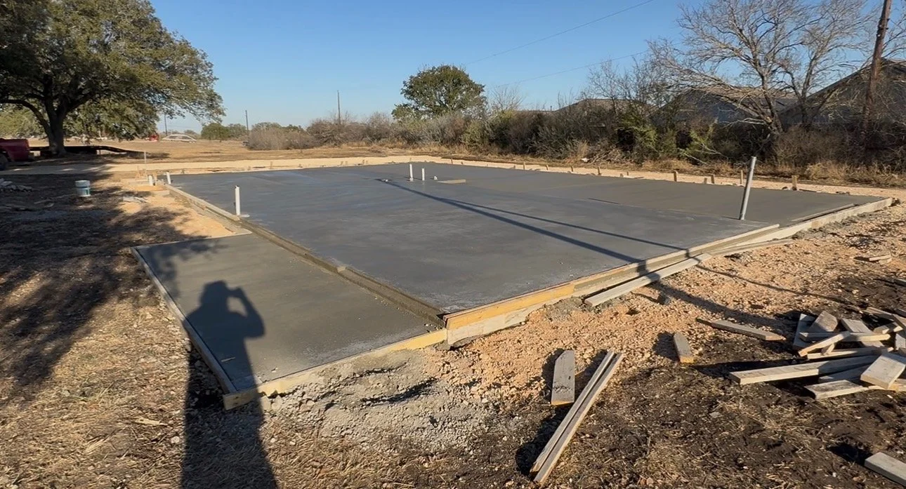 Freshly poured concrete foundation on a construction site with wooden framing, pipes for utilities, and trees in the background.