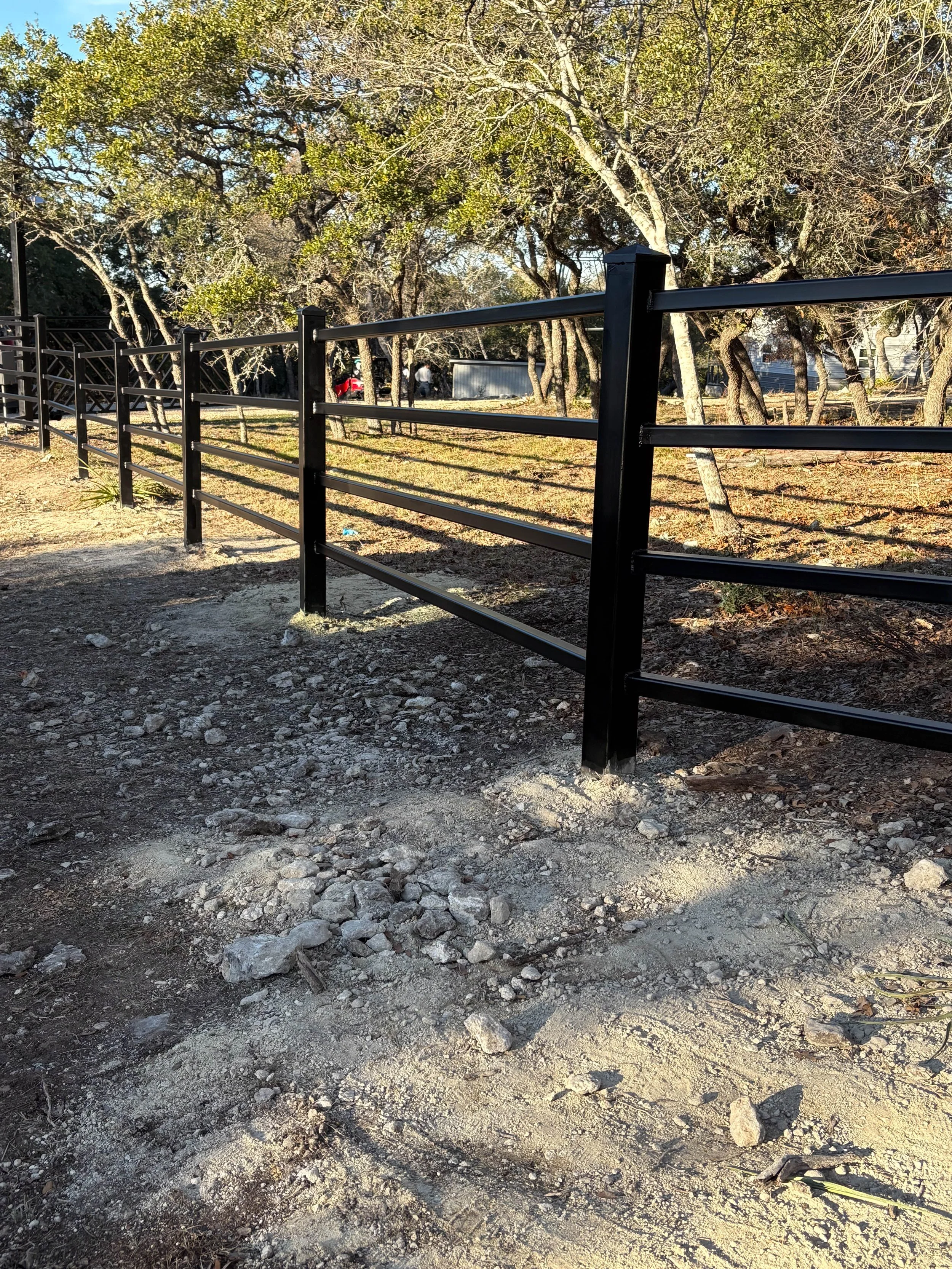 A black metal fence along a dirt path with gravel and small rocks, behind which there are trees with green and brown leaves and a small shed in a backyard.
