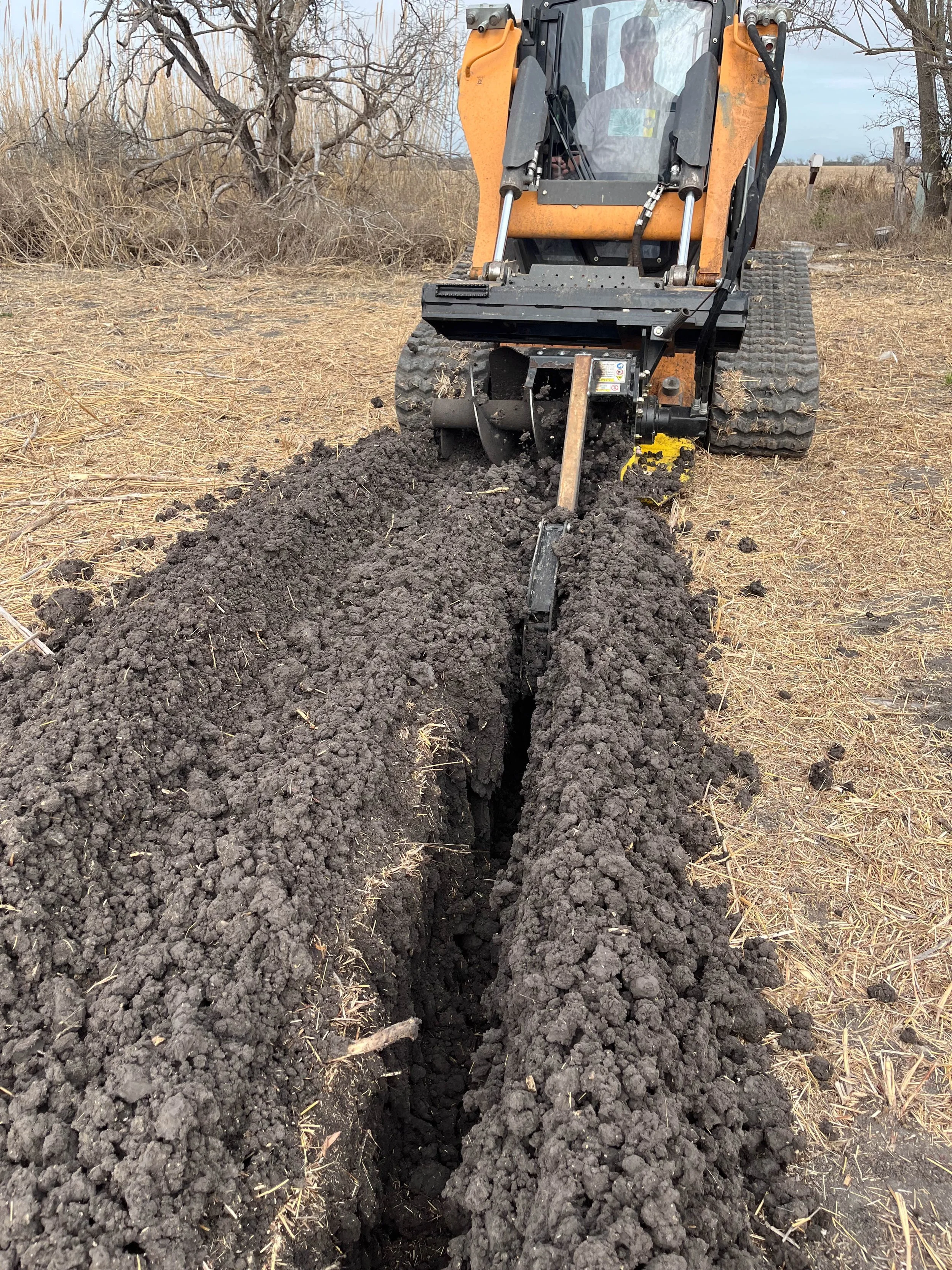 A small orange excavator digging a narrow trench in a rural outdoor area with dry grass and leafless trees in the background.