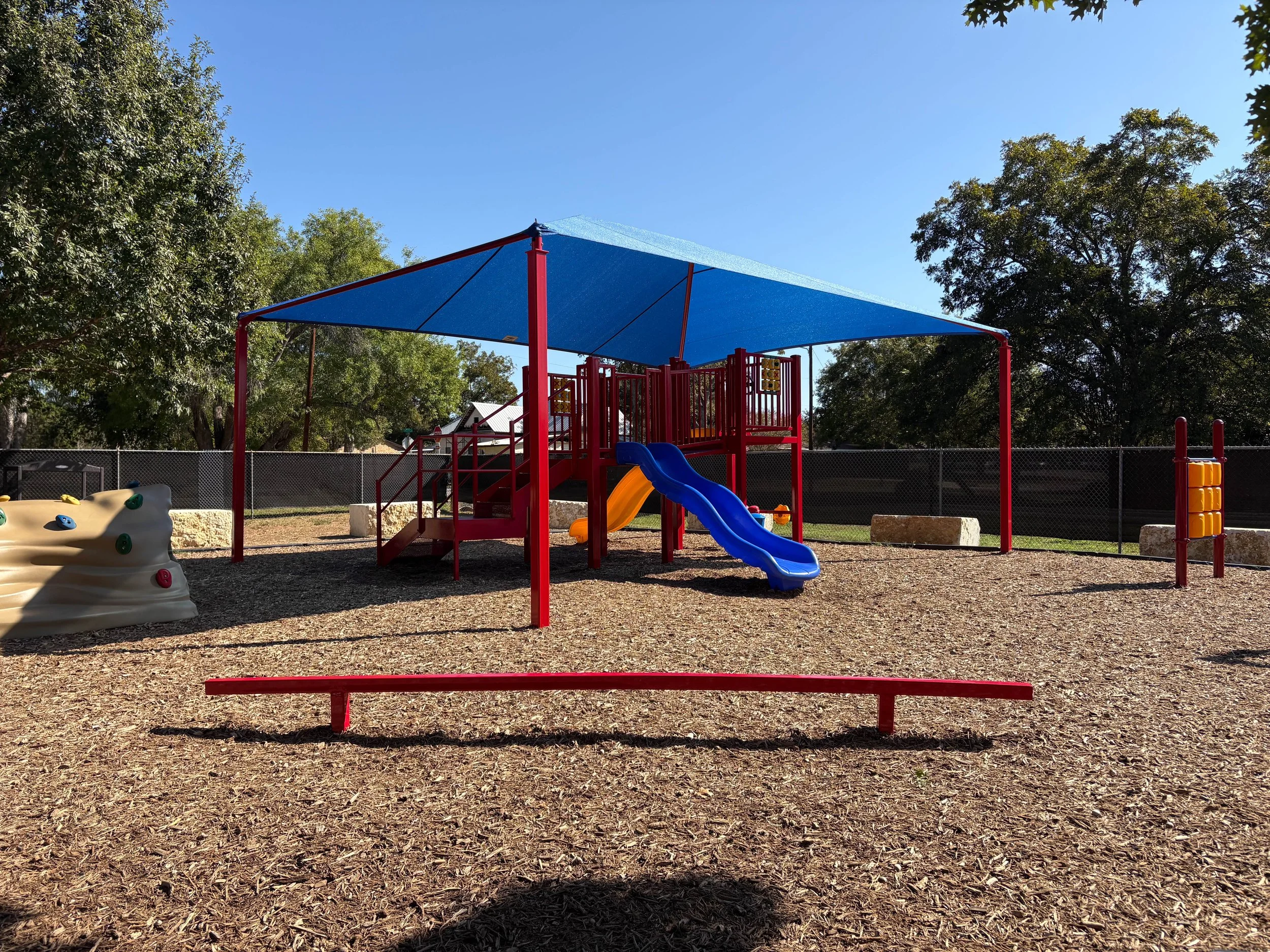 Colorful playground with a red and blue covered play structure, a small slide, rock climbing wall, and various play equipment on a wood chip surface, surrounded by trees under a clear blue sky.