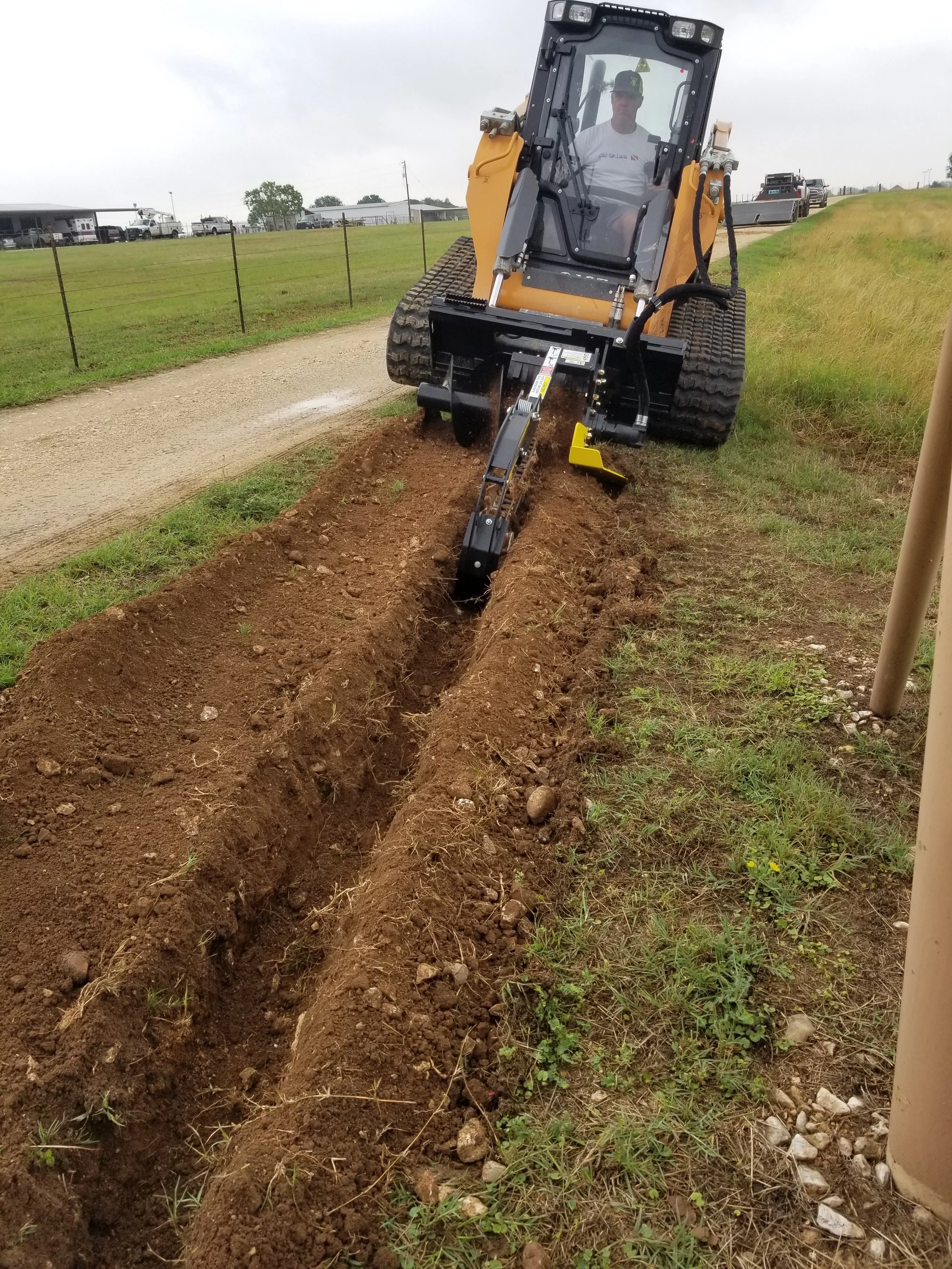 A person operating a small excavator digging a trench on the side of a dirt path. The operator is visible inside the cabin, and several vehicles are parked on a hill in the background.