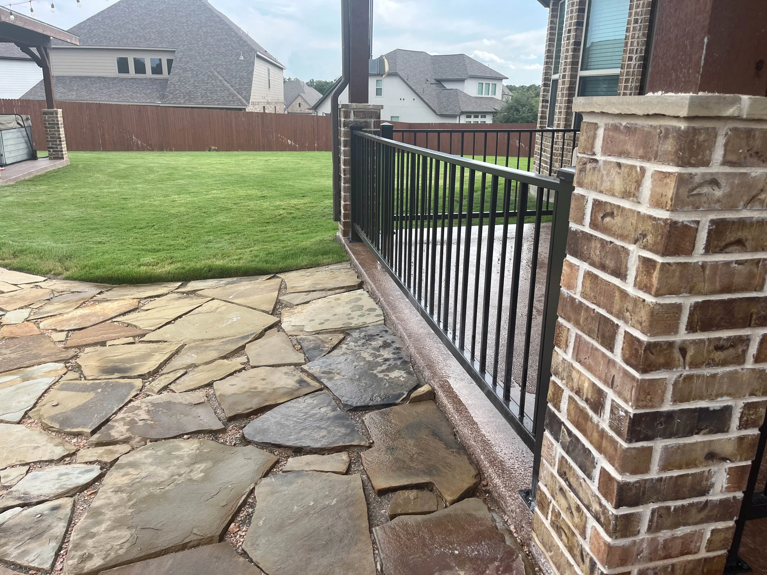 View of a backyard with green grass, a stone patio, a brick house wall, and a wooden fenced yard with neighboring houses in the background.
