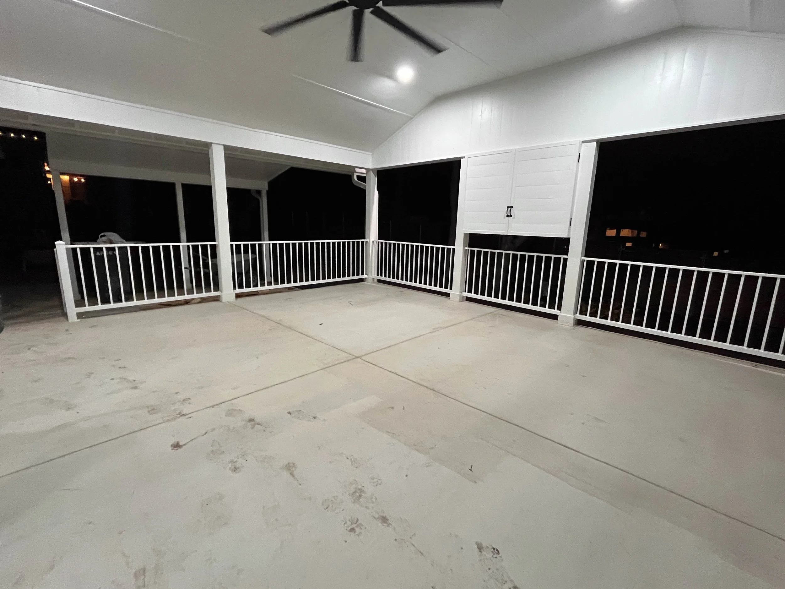 Nighttime view of a porch with a white railing, a ceiling fan, and a ceiling light, with footprints visible on the concrete floor.