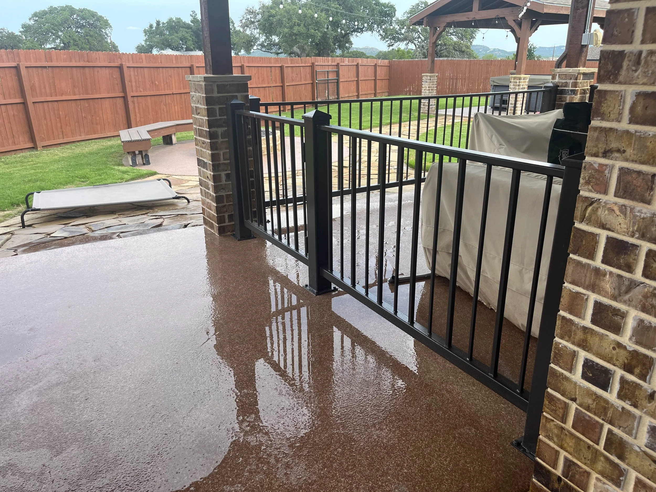 Backyard patio scene with a black metal gate, a fenced yard, and a covered patio area with string lights, a grill covered with a beige cover, and a stone pathway, after rain.