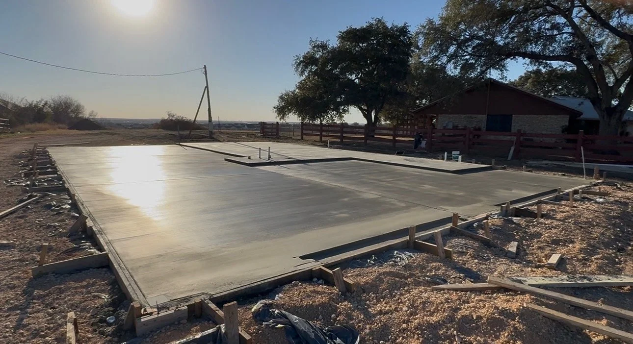Freshly poured concrete slab for a building foundation on a construction site, with wooden formwork and rebar, under a clear sky with the sun visible in the background.