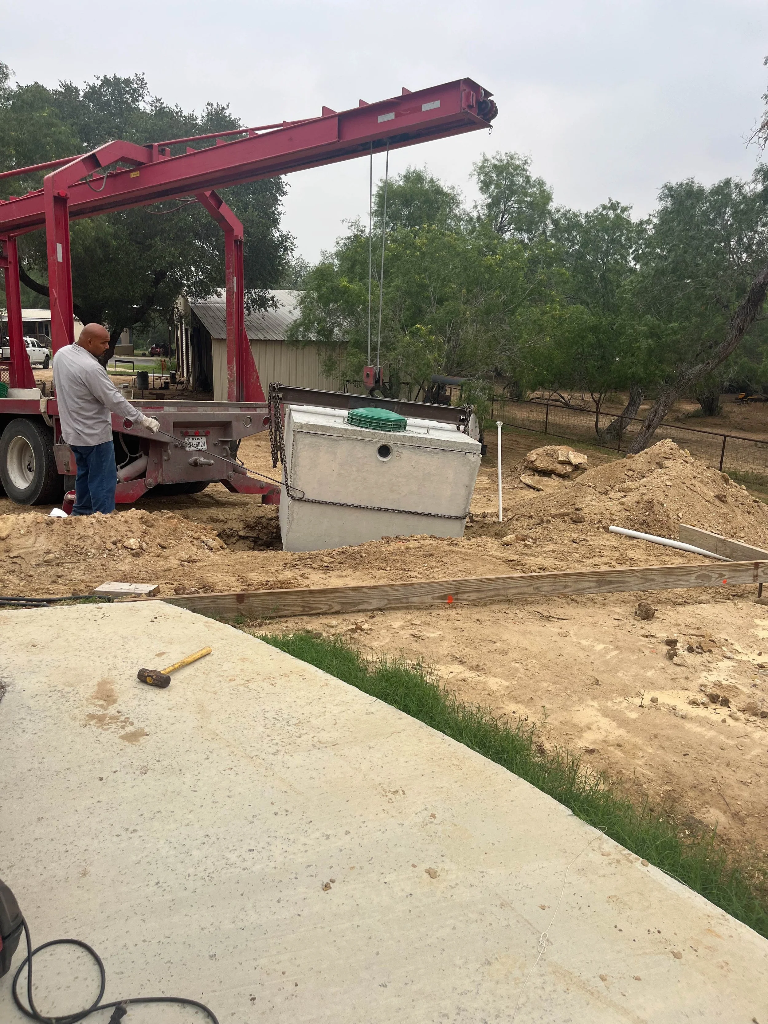 Construction site with a man in a light-colored shirt and jeans working near a concrete structure, a red crane, dirt, and trees in the background.
