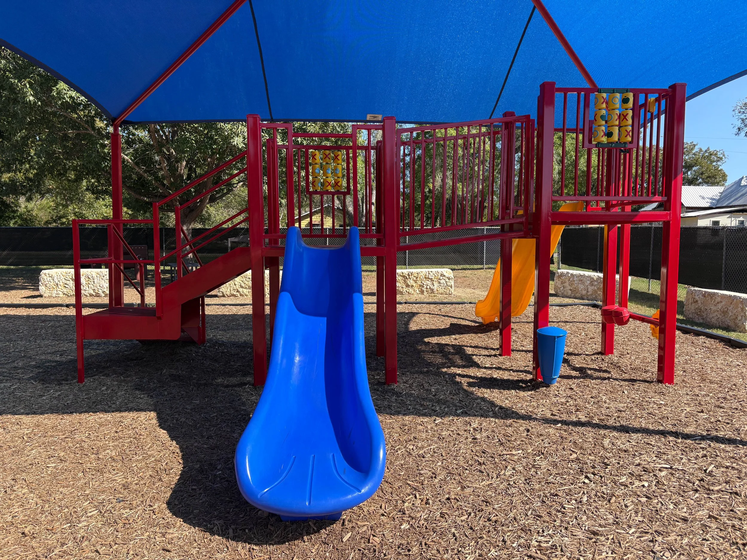 Colorful playground with blue slide, yellow slide, and tic-tac-toe game, shaded by a large blue canopy, in a park with trees and houses in the background.