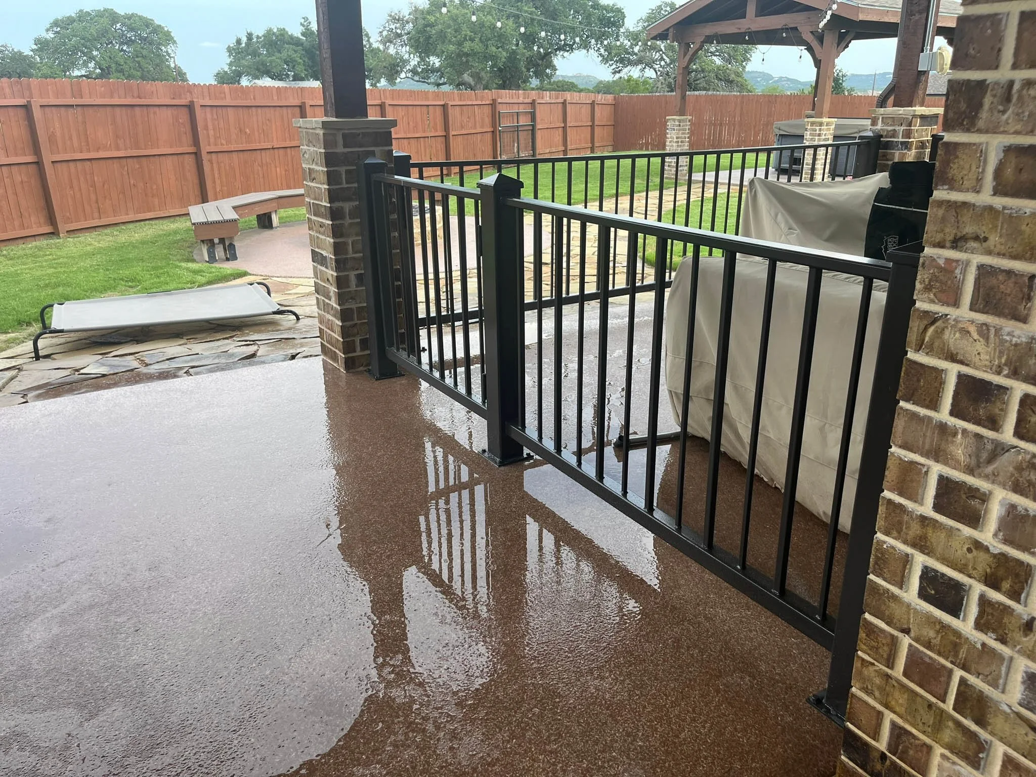 A backyard patio with a wet concrete surface, a golden retriever lying on a white mat, a black fence, brick pillars supporting a wooden pergola, and a wooden fence surrounding the yard.