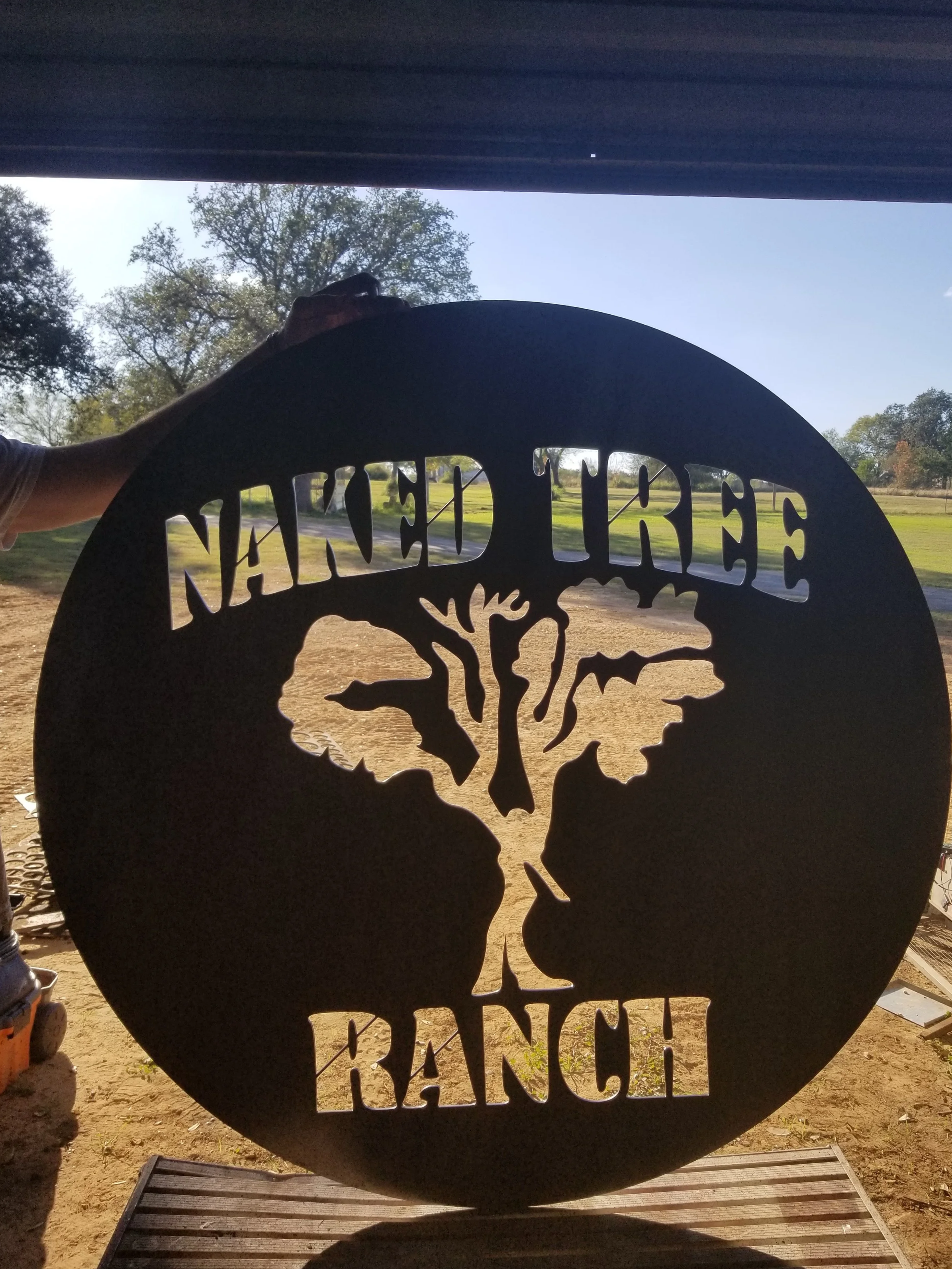 Metal sign shaped like a skull with the words "Never Tread" at the top and "BANG!" at the bottom, outdoors with trees and a clear sky in the background.