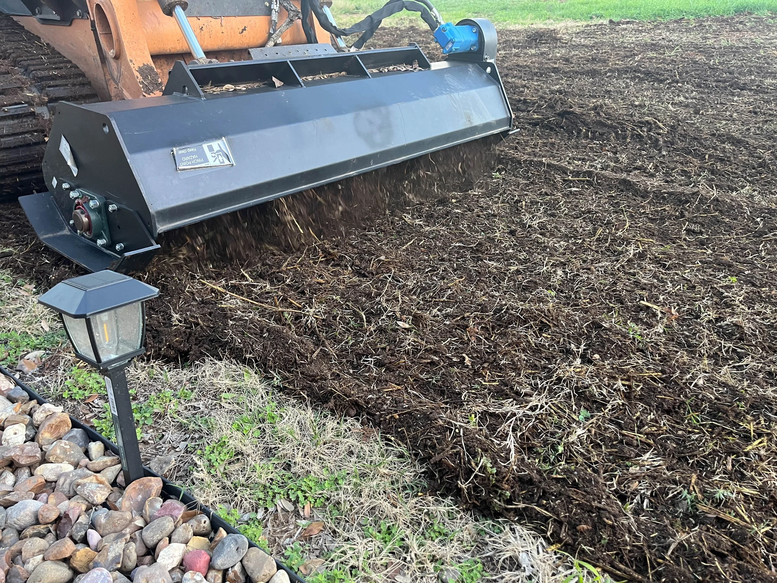 A large industrial soil tiller attachment connected to a construction vehicle, used for tilling soil in a garden or landscape. The attachment is rotating, breaking up and mixing the soil, with a mix of dirt and plant debris visible. There is a pathwa