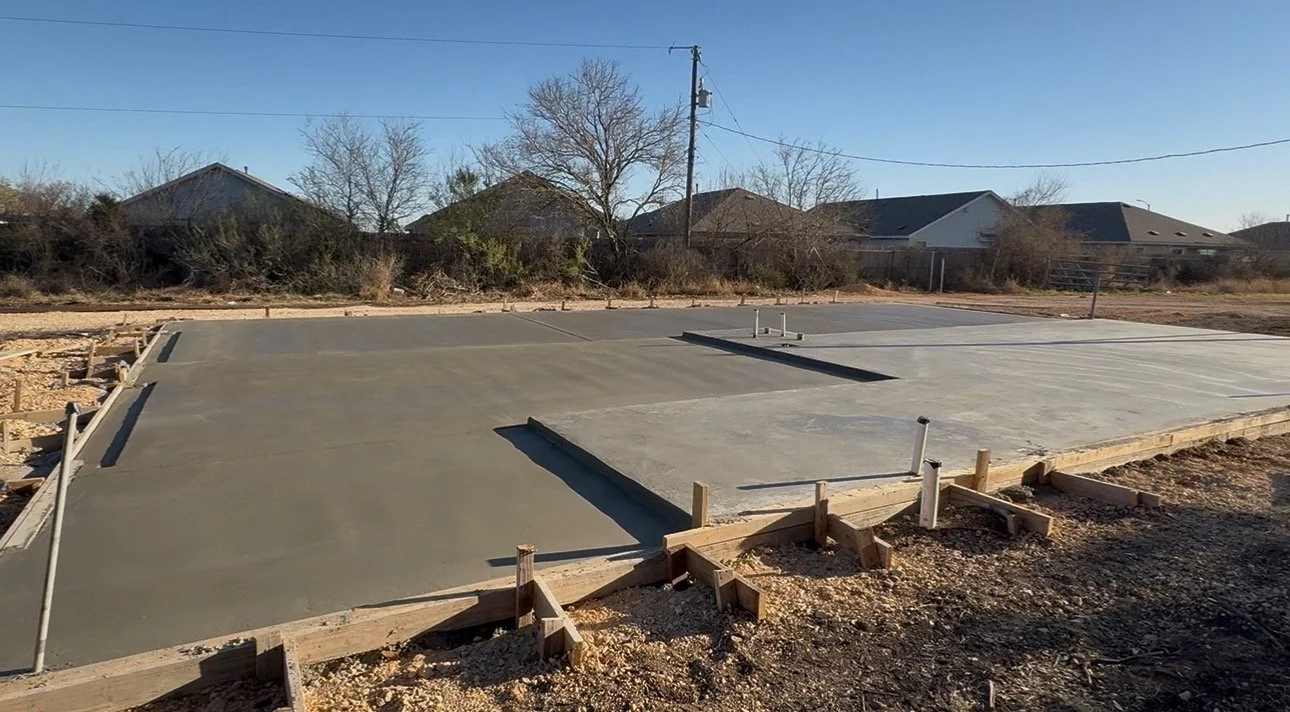 Concrete foundation poured for a building with wooden formwork and pipes protruding, on a construction site with houses and trees in the background.