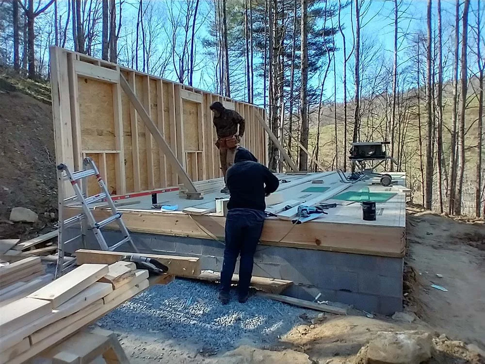 Two people working on framing a house in a wooded area. One is standing on the ground, the other is on the platform, with construction tools and materials scattered around.
