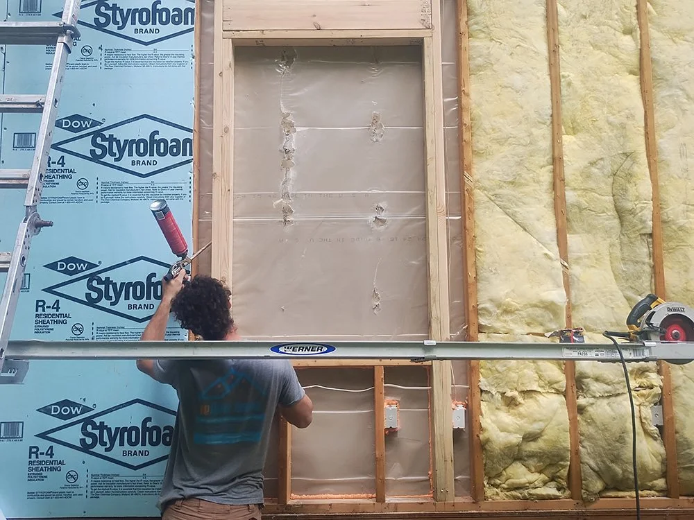A construction worker installing or working on insulation in a building wall with wooden framing. The wall has blue foam insulation on the left and yellow fiberglass insulation on the right. The worker is holding a red caulking gun, and there is a ladder on the left side of the image.