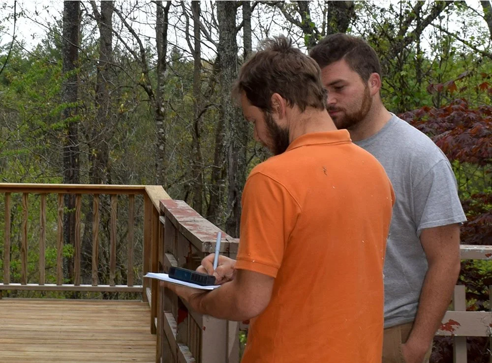 Two young men, one in an orange shirt with a beard, and the other in a gray shirt, are standing on a wooden deck outdoors. The man in orange is writing on a notepad, while the other looks over. They are surrounded by trees and foliage.