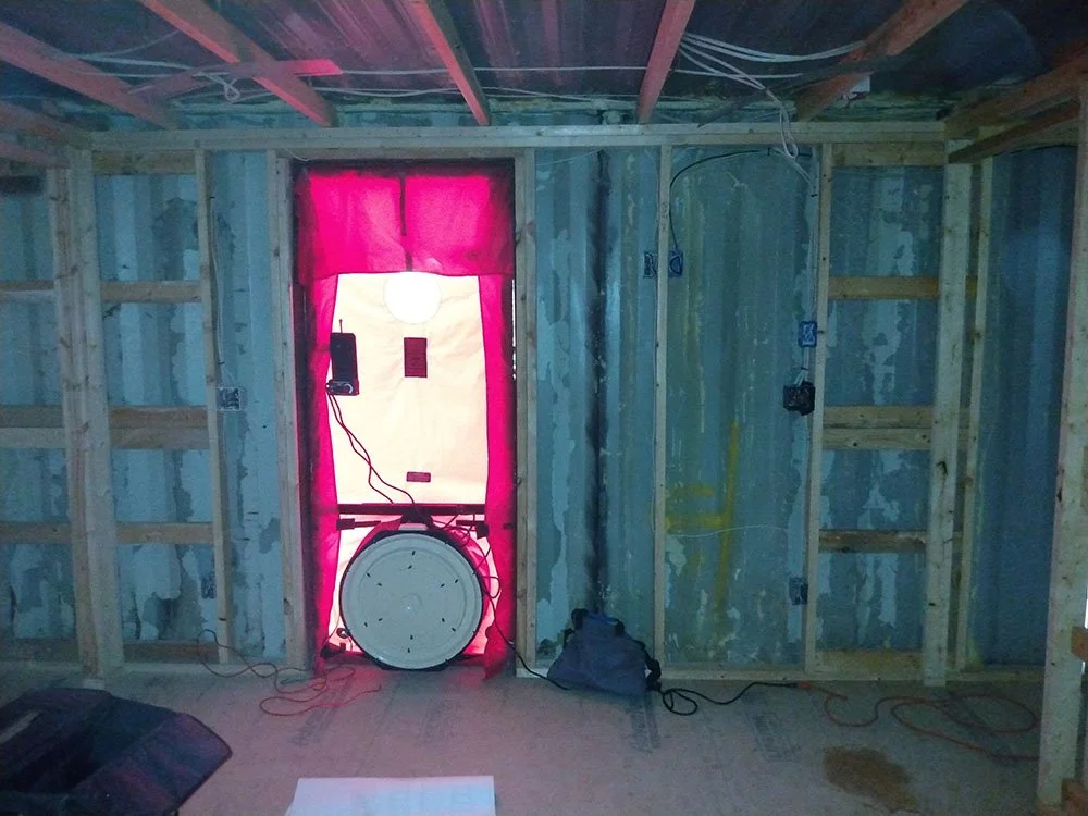 Construction room with uninsulated wooden framing, wiring, and a window covered with pink material. There are electrical boxes and a blower door installed for testing.