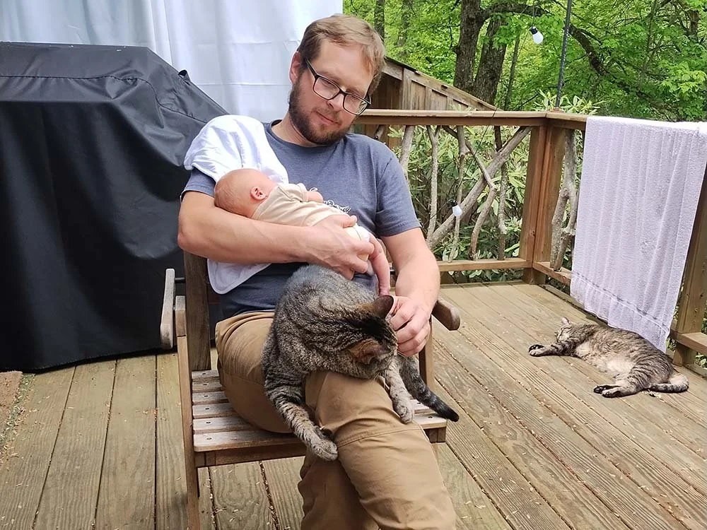 A man holding a baby and sitting on a wooden deck with a tabby cat on his lap; another cat lying on the deck nearby, outdoors in a green wooded area.