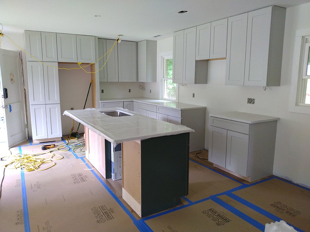 Kitchen under construction with white cabinets, a central island with a countertop, and unfinished flooring with blue painter's tape.