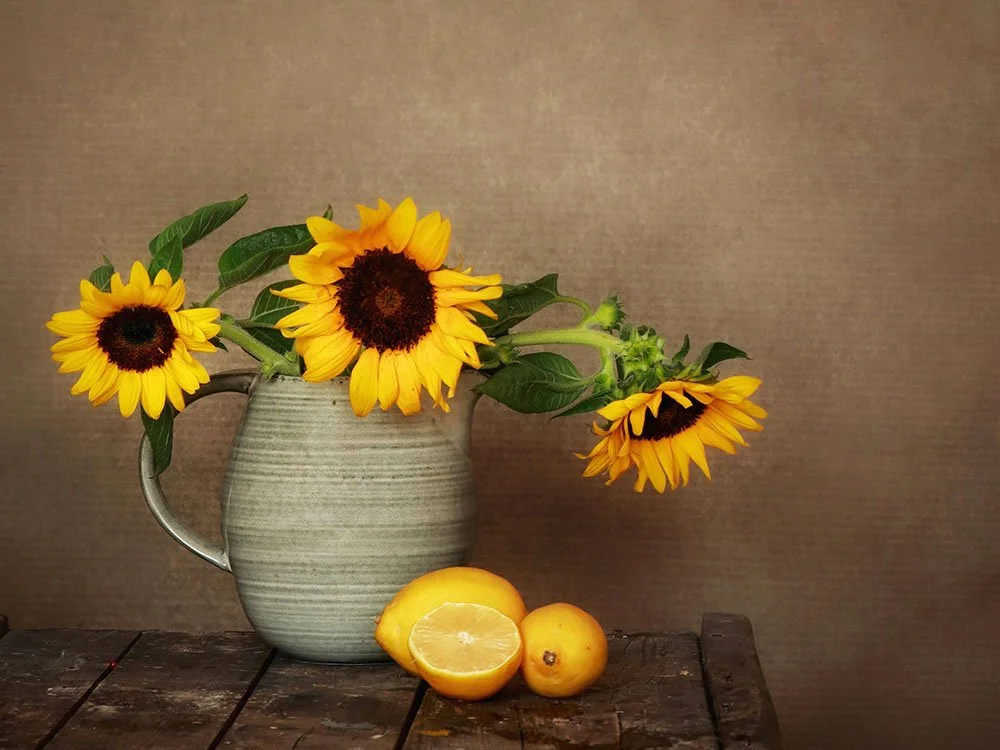 Sunflowers in a gray ceramic pitcher with lemons and lemon halves on a wooden table against a neutral background.