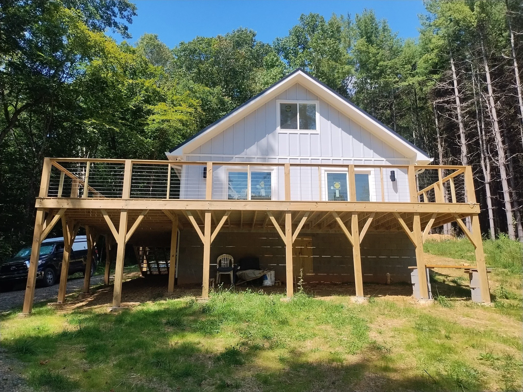 A house under construction with an elevated deck, surrounded by trees and parked cars, on a sunny day.