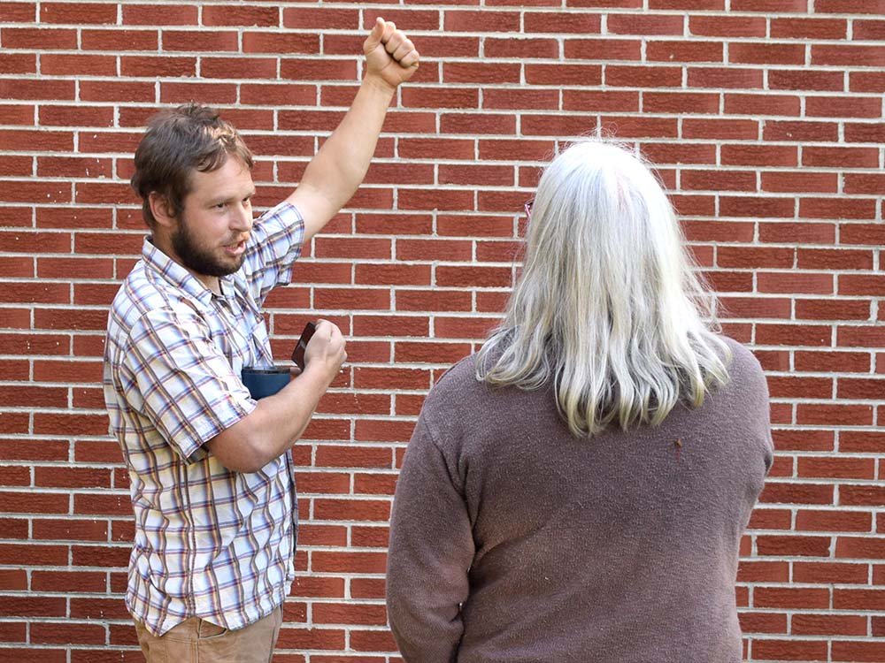 A man with a beard and plaid shirt raising his fist and talking to a woman with long gray hair, standing in front of a brick wall.