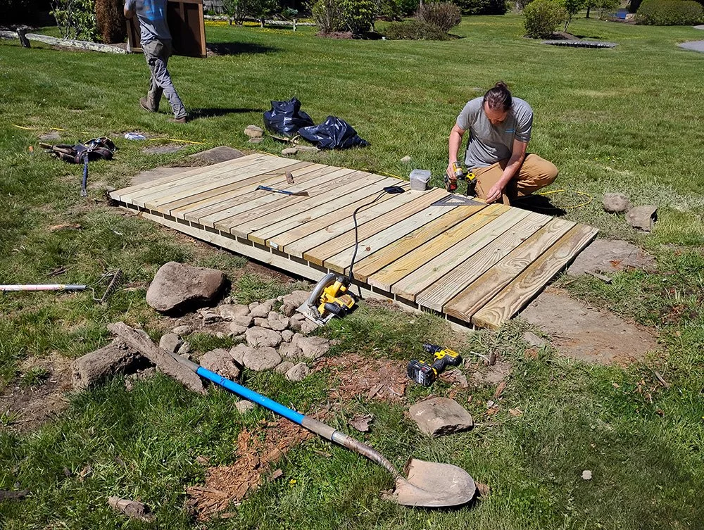Two people constructing a wooden deck on a grassy lawn, with tools and materials scattered around.