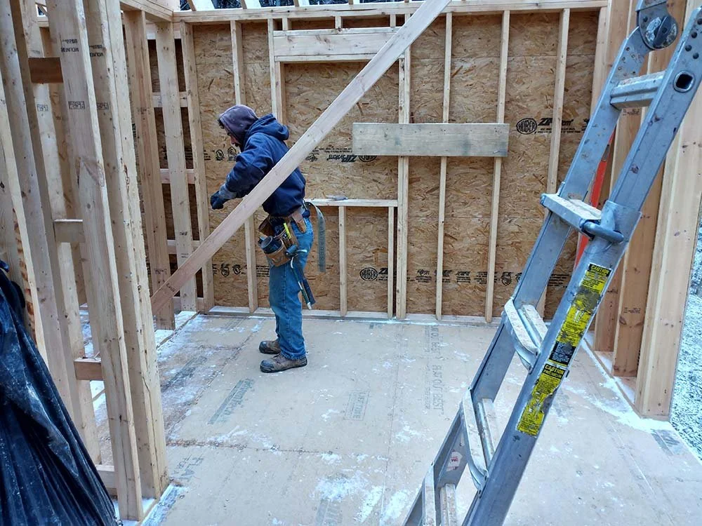 A construction worker in blue clothing and a hoodie working inside a partially framed wooden structure during building.