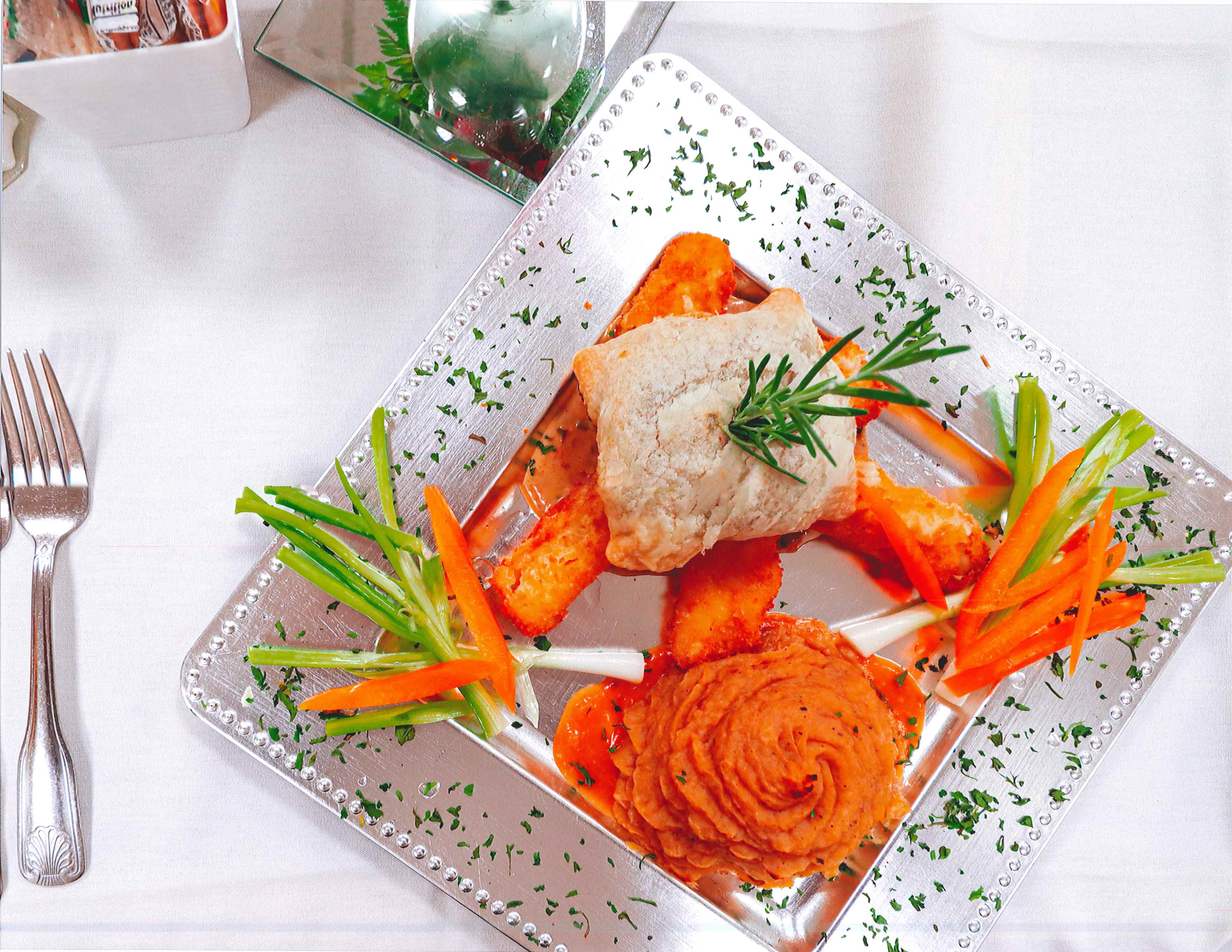A silver tray with fried fish, roasted carrots, and green veggie garnishes, served alongside a white tablecloth.