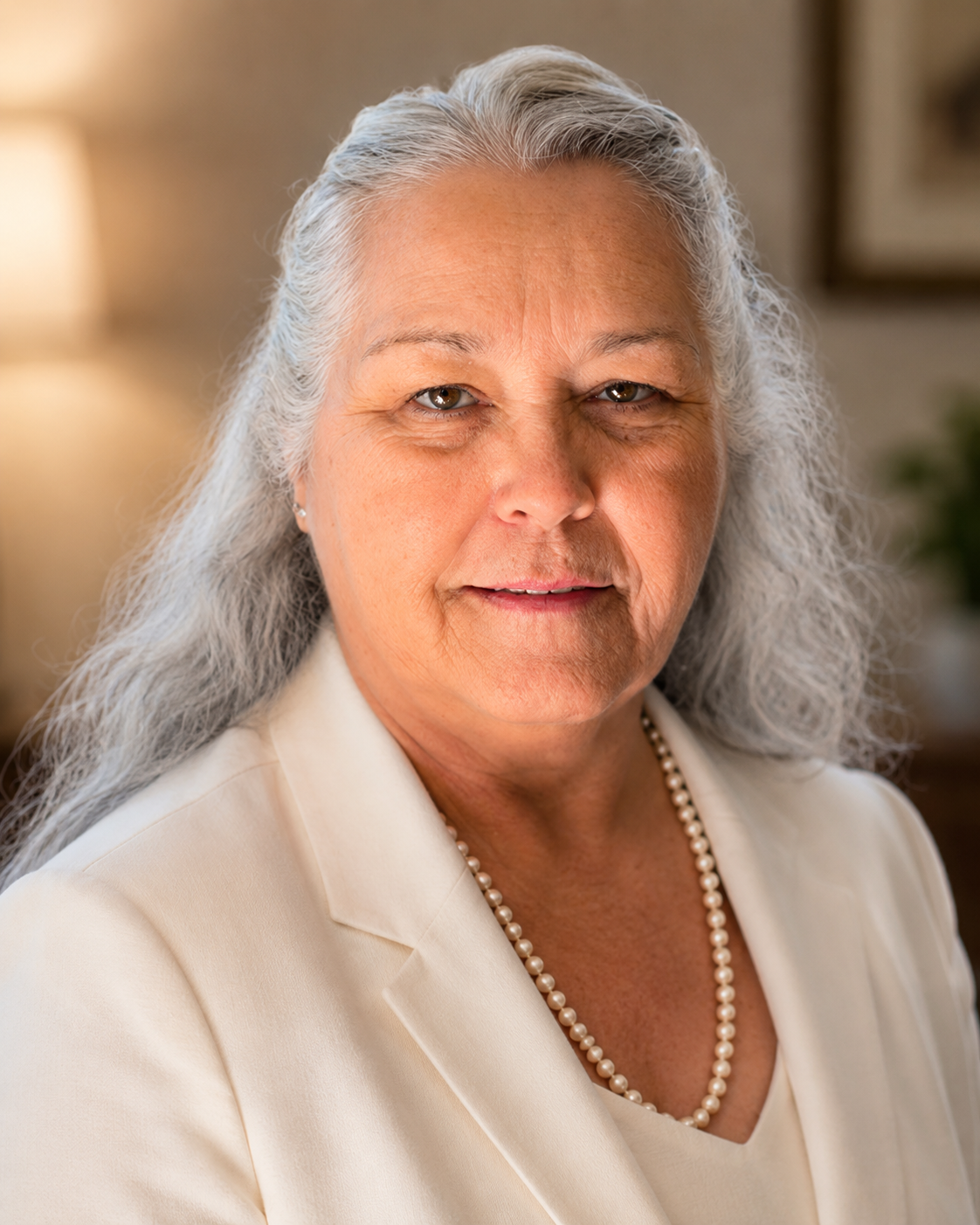A senior woman with silver hair wearing a cream-colored suit, pearl necklace, and earrings, looking at the camera with a warm expression in a well-lit home setting.