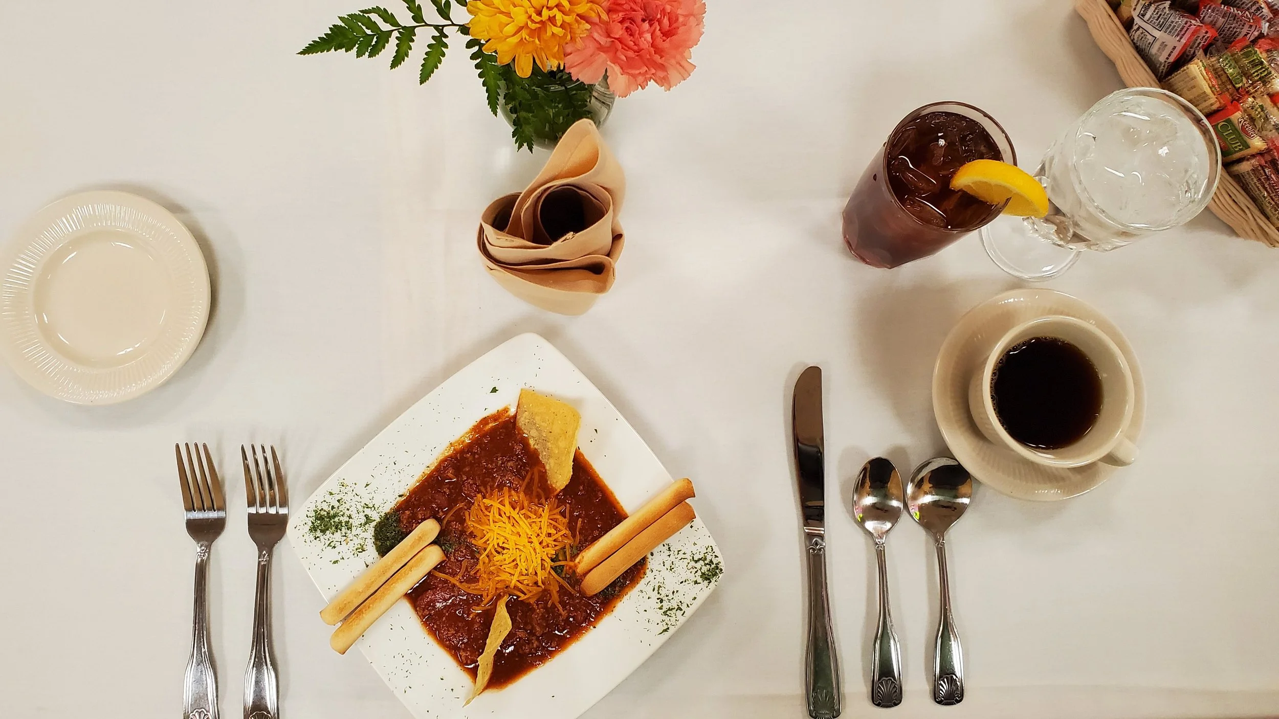 A dining table set with utensils, a plate of spicy Mexican beef stew with cheese, a glass of iced tea with lemon, a cup of coffee, and a flower arrangement.