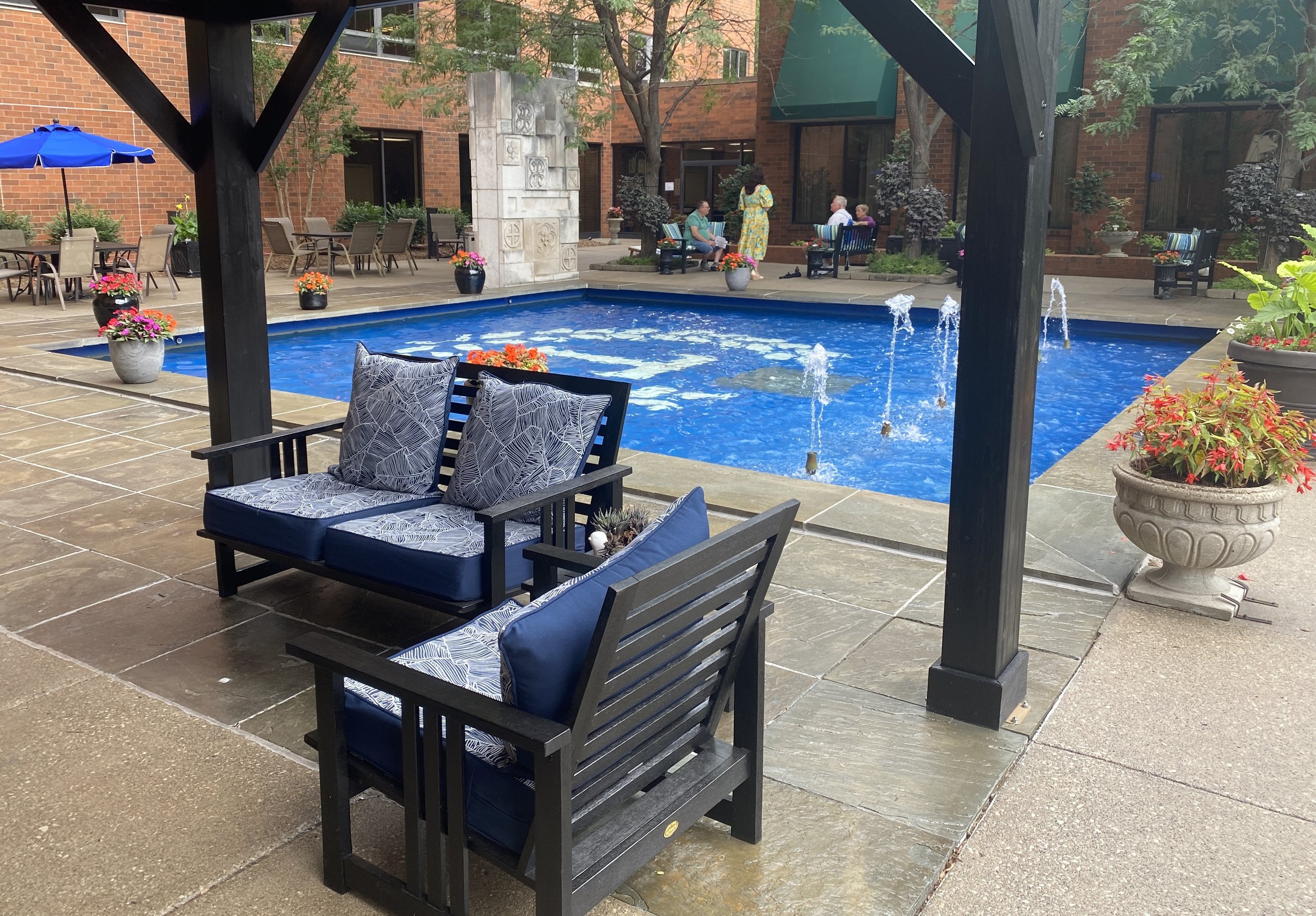 An outdoor patio area with a swimming pool, surrounded by chairs, tables, potted plants with flowers, and people sitting and socializing.