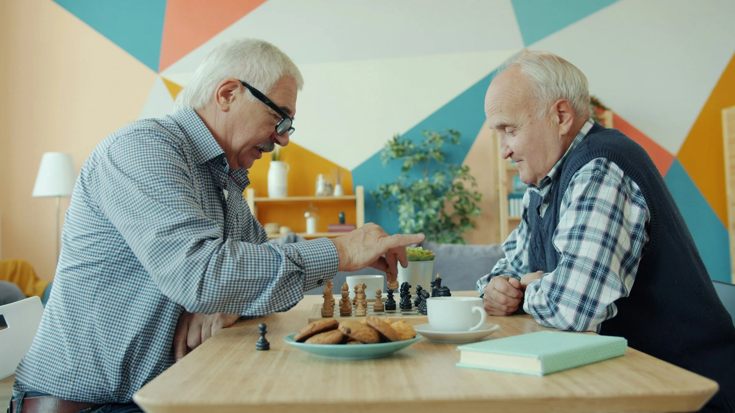Two elderly men playing chess at a wooden table, with cookies and a book nearby, in a colorful room.