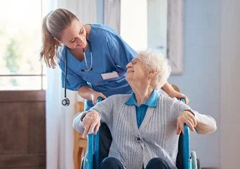 A healthcare worker smiling at an elderly woman in a wheelchair inside a bright room.
