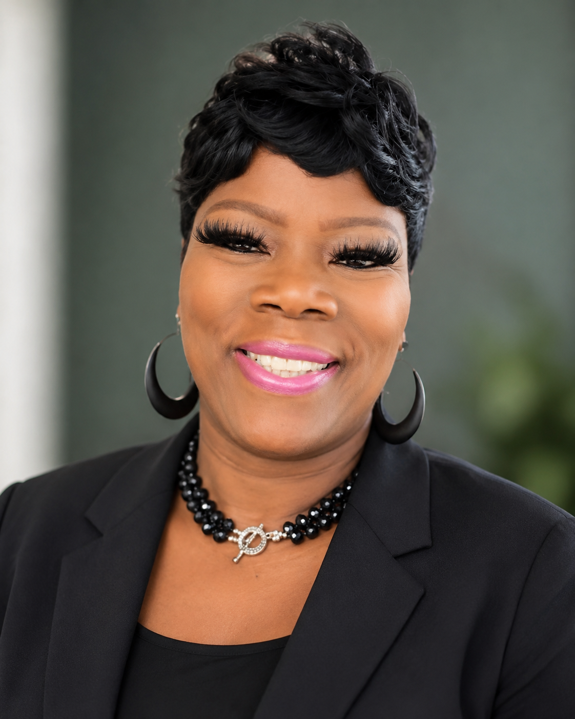 Close-up portrait of an African American woman with short black hair, bright pink lipstick, and large black hoop earrings, wearing a black blazer and a black beaded necklace with a silver 'P' pendant.