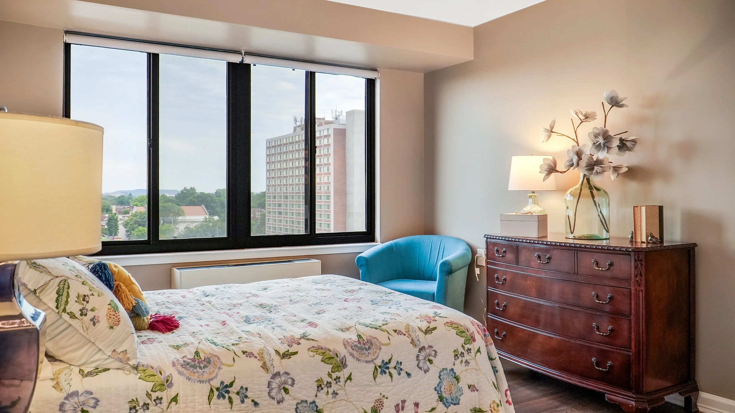 Interior of a bedroom with a large window showing a cityscape, a bed with a floral quilt, a wooden dresser with a lamp, a glass vase with flowers, and a blue armchair.