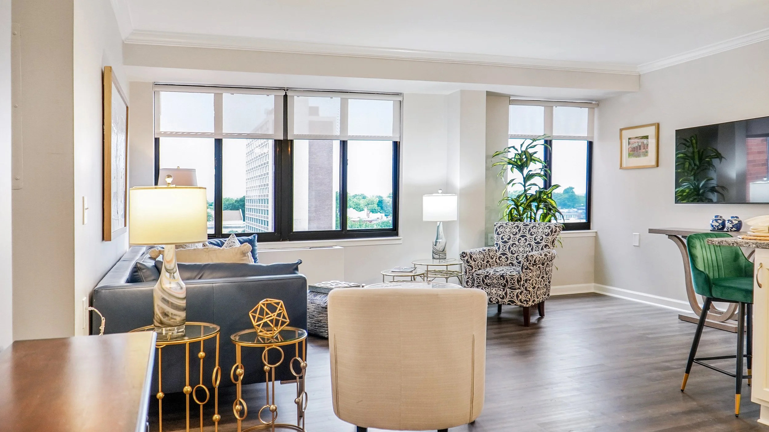 Living room with large windows, modern furniture, a patterned armchair, a black leather sofa, a glass coffee table, and indoor plants.